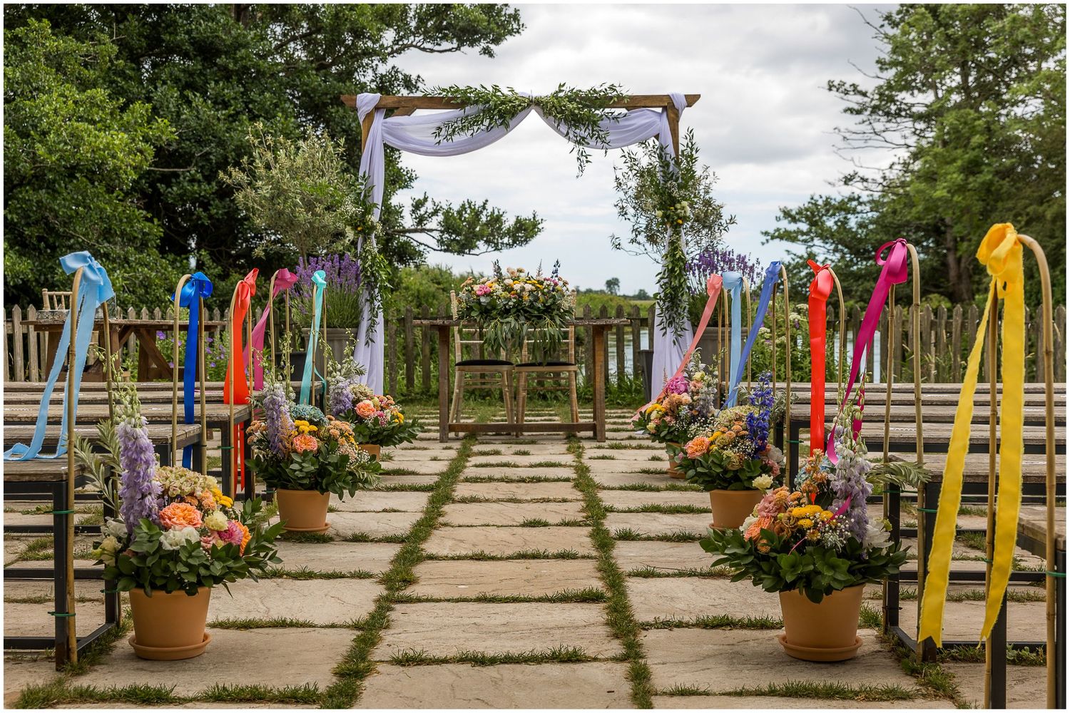 Colorful outdoor wedding ceremony aisle with rainbow ribbons, floral arrangements in terracotta pots, and a decorated arch backdrop