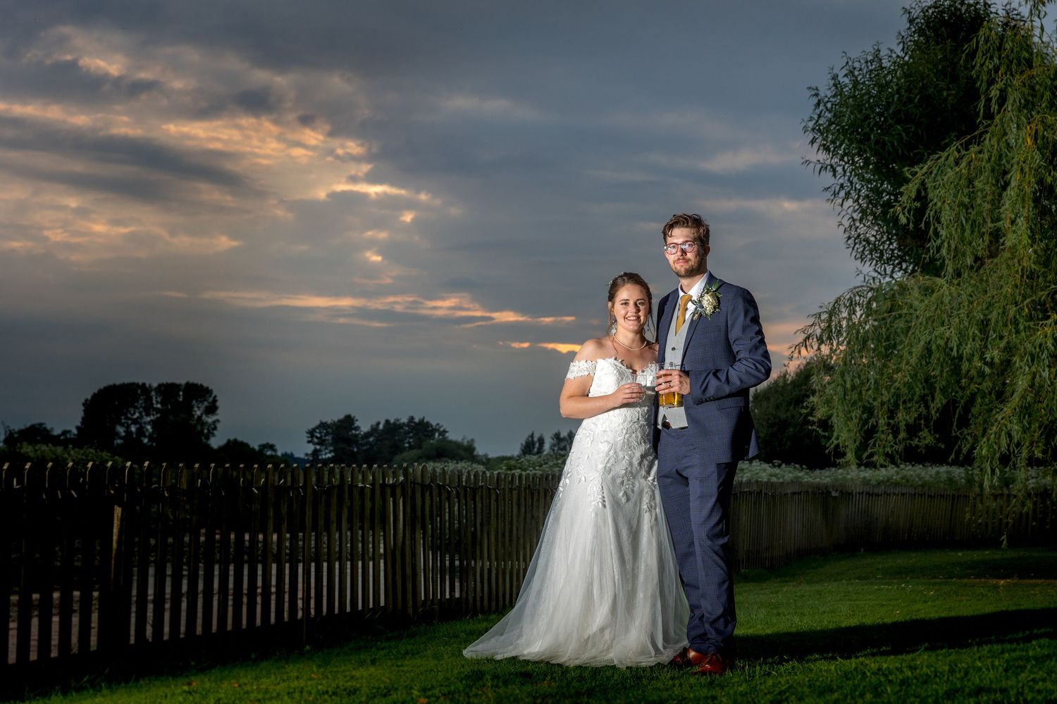 Dramatic sunset silhouettes a couple against a moody sky with trees and fence in the background.