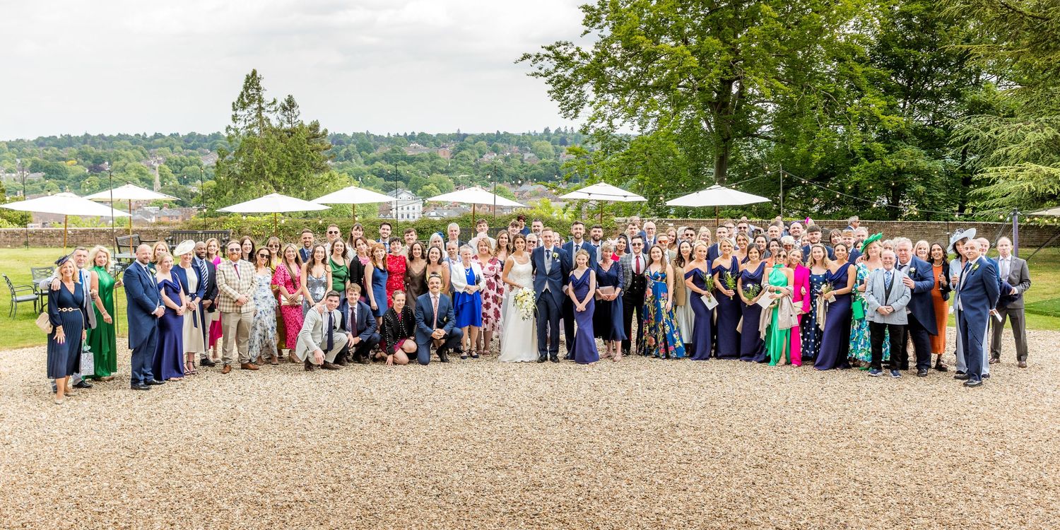 Large group wedding photo taken outdoors with scenic countryside backdrop.