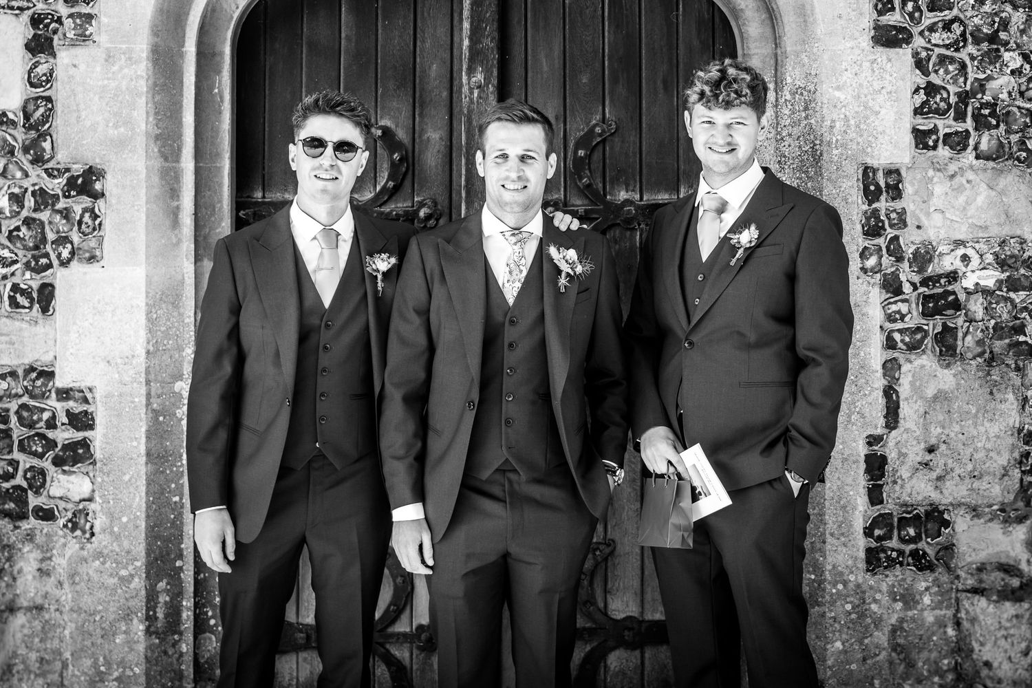 Groomsmen in black suits gather for formal wedding photos outside a historic stone building.