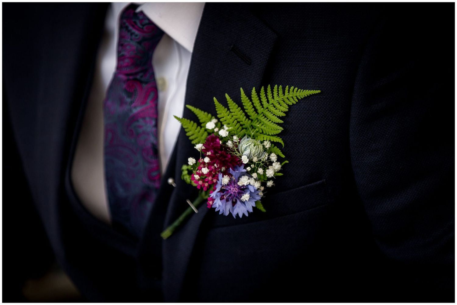 A formal boutonniere with purple and white flowers and green fern leaves adorns a dark suit lapel.