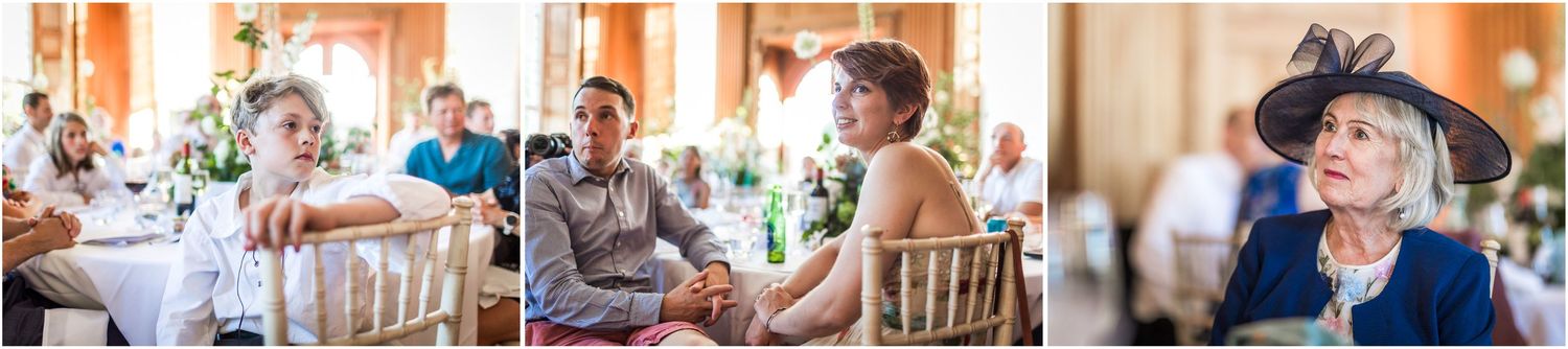 Guests seated at wedding reception tables, smiling and chatting, with an elegantly dressed woman in a blue hat in the foreground.