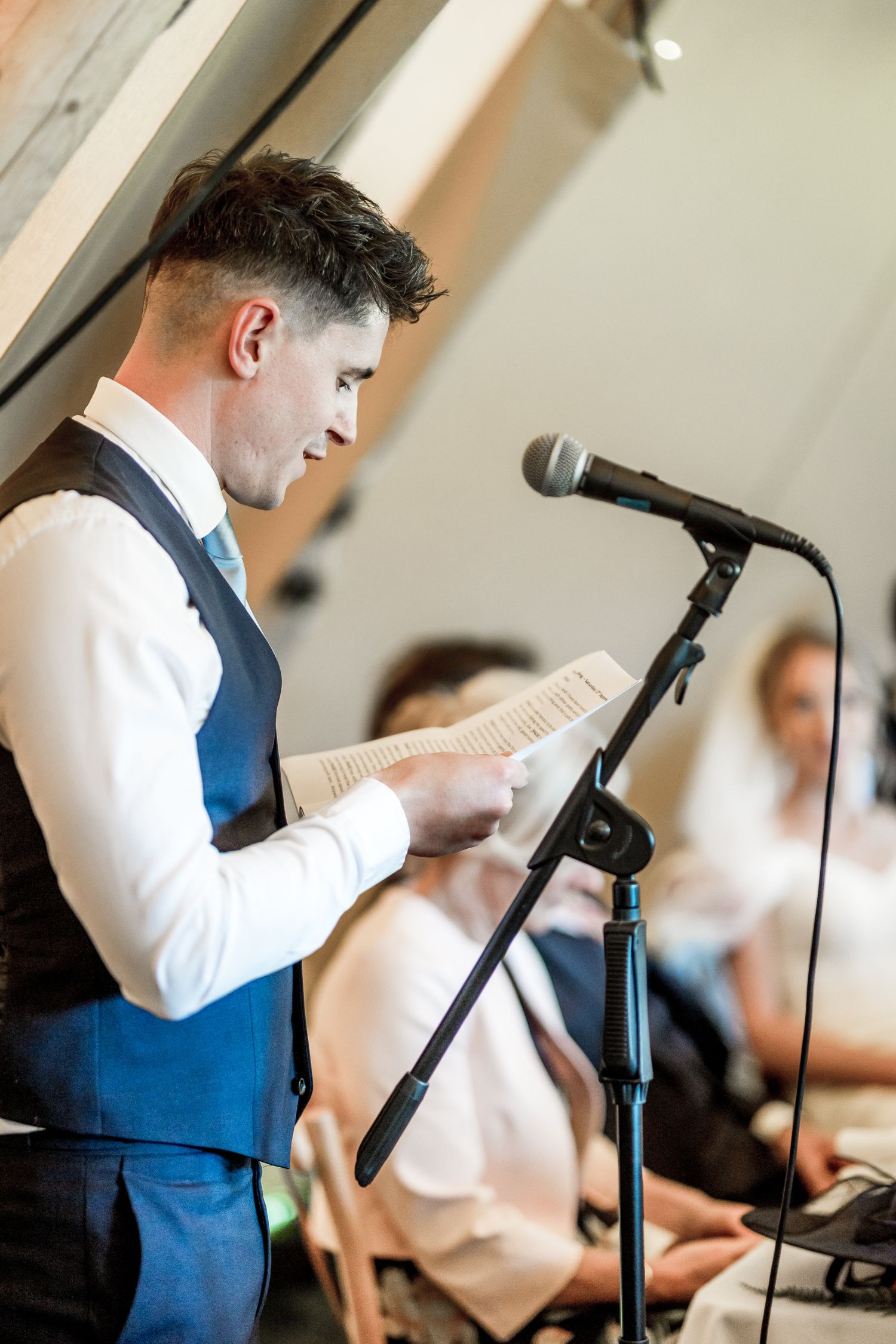 Speaker at microphone delivers wedding speech wearing blue vest and white shirt.