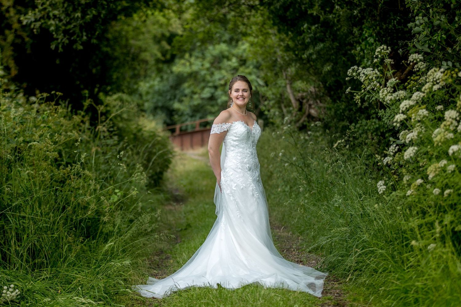 Elegant bride in lace dress poses among flowering bushes and greenery.