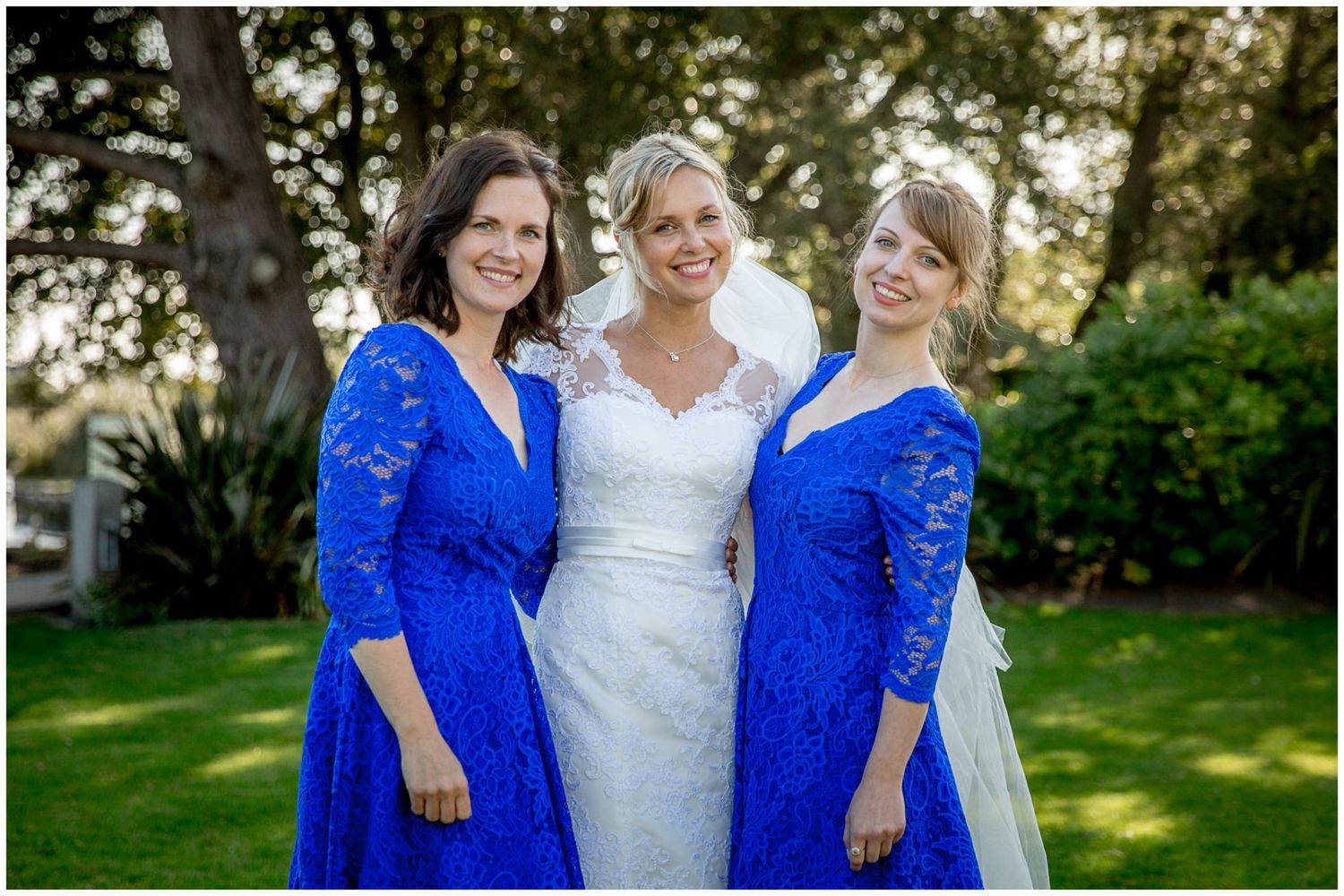 Three women pose together outdoors wearing matching royal blue lace dresses and a white wedding gown in a garden setting.