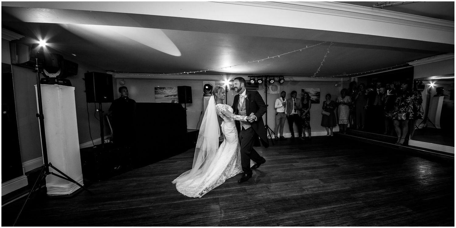 A black and white wedding reception photo showing dancing guests on a dark dance floor with dramatic lighting.