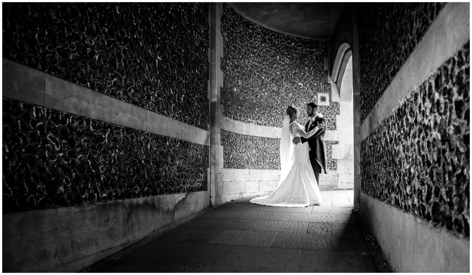 A black and white wedding photo captures a couple in a dramatic backlit tunnel with textured walls.
