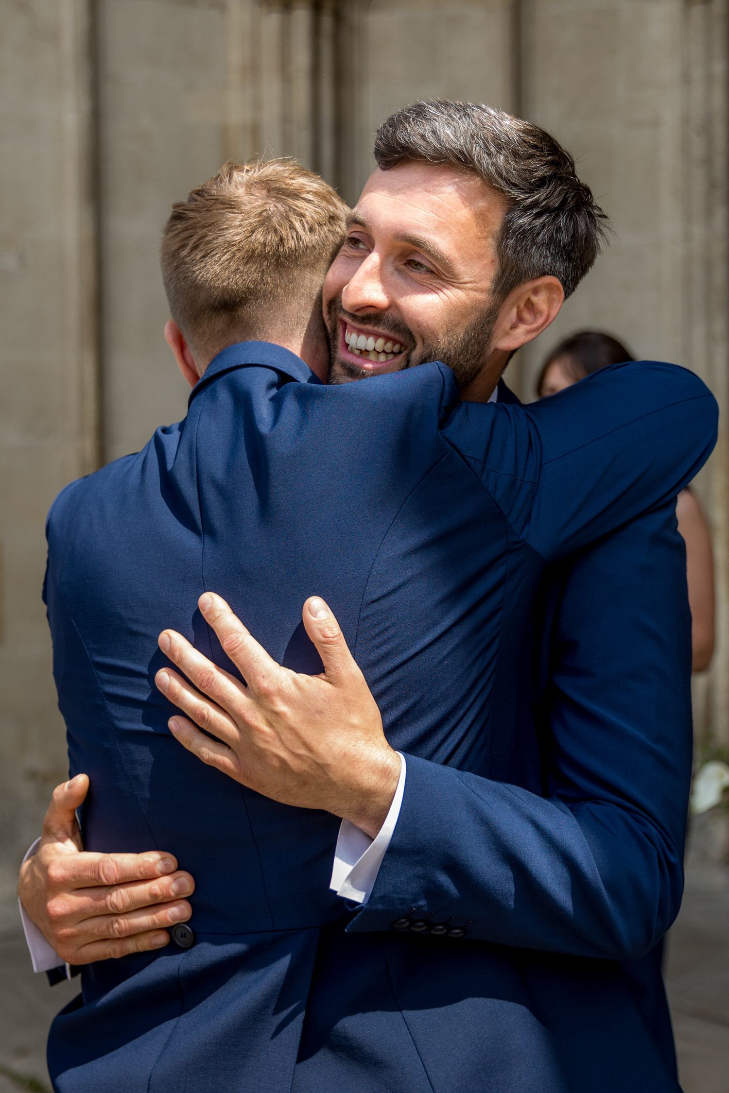 Emotional embrace between two people wearing navy blue suits during a wedding celebration.