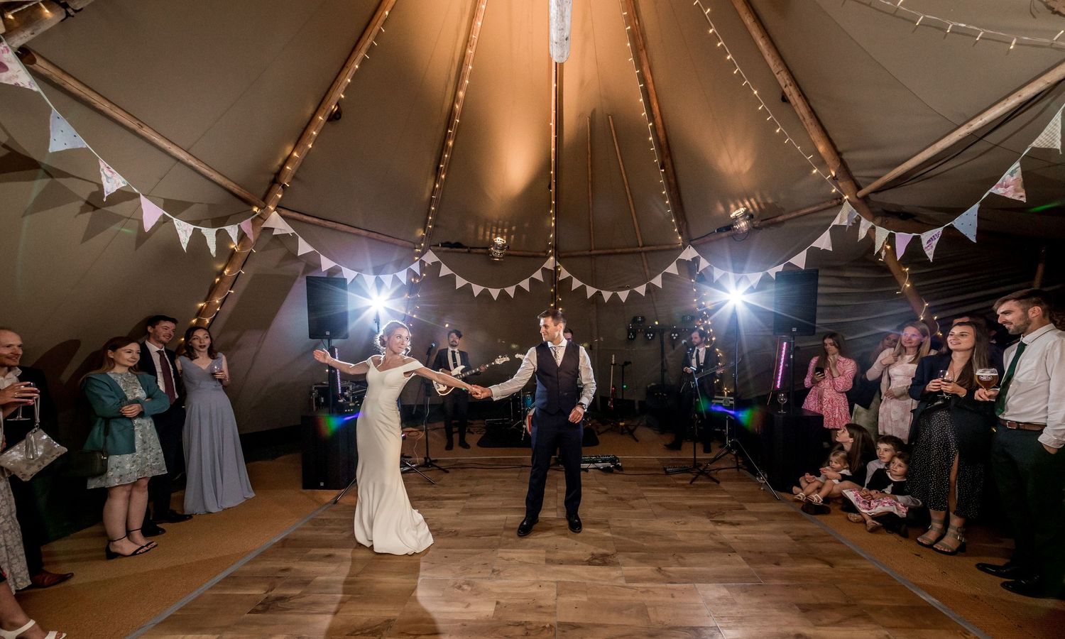 A newlywed couple dances under string lights in a wedding tent while guests watch from the sides.
