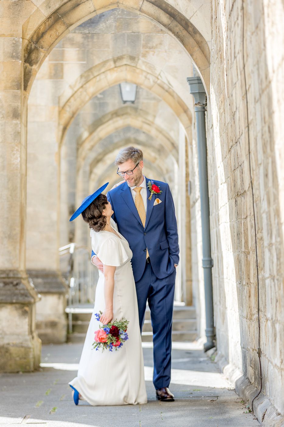 Wedding couple in formal attire walking through stone archways in bright natural light.