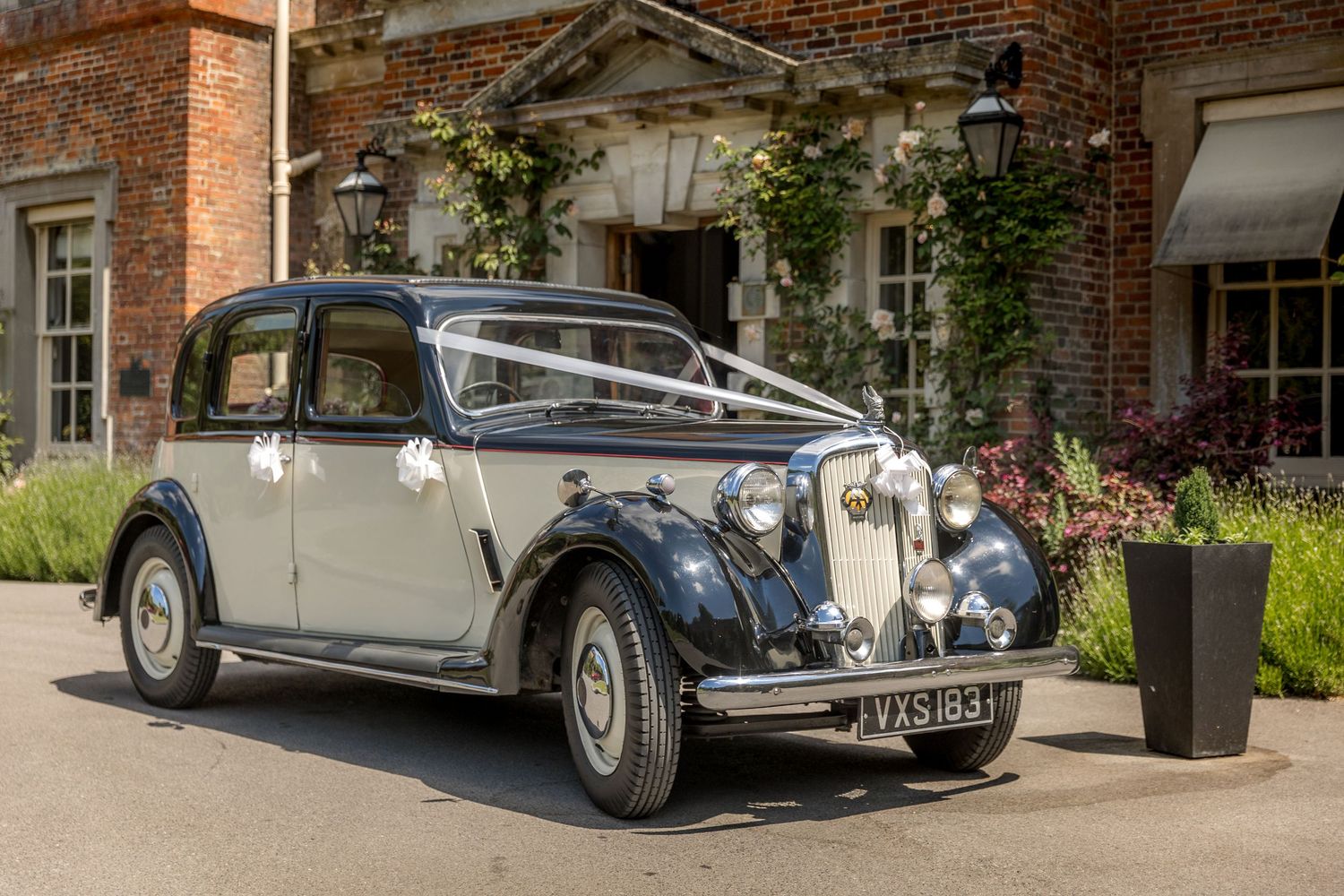 Classic vintage cars parked outside a historic brick manor house on a sunny day.