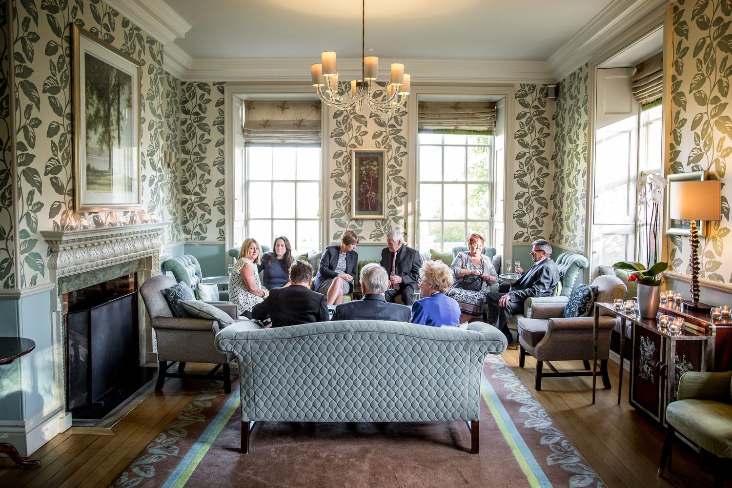 Guests seated in an elegant drawing room with floral wallpaper and chandelier during a wedding event.
