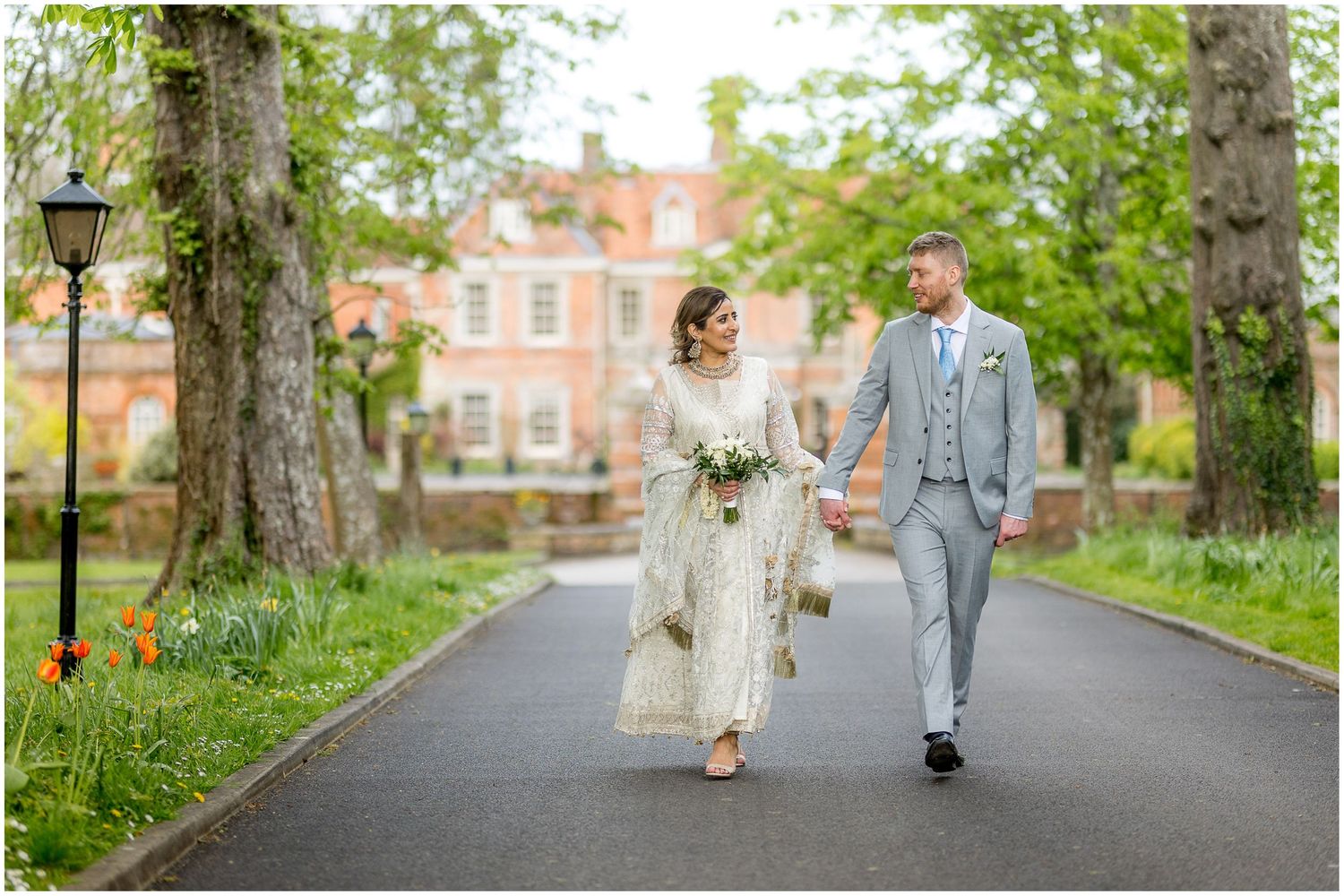 The bride and groom walk down the driveway at Lainston House Hotel with the wedding venue in the background