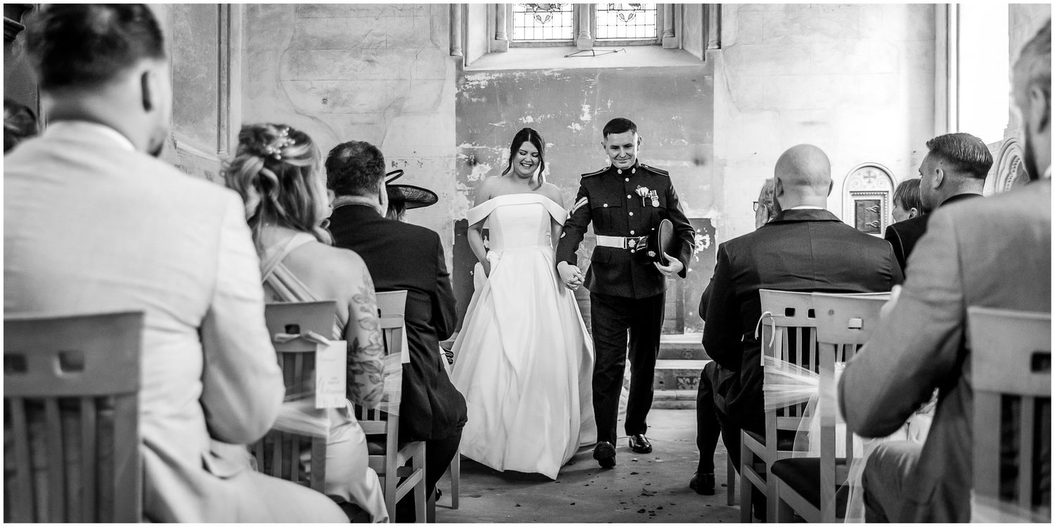 A couple walks down the church aisle at The Elvetham while guests look on during a wedding ceremony.