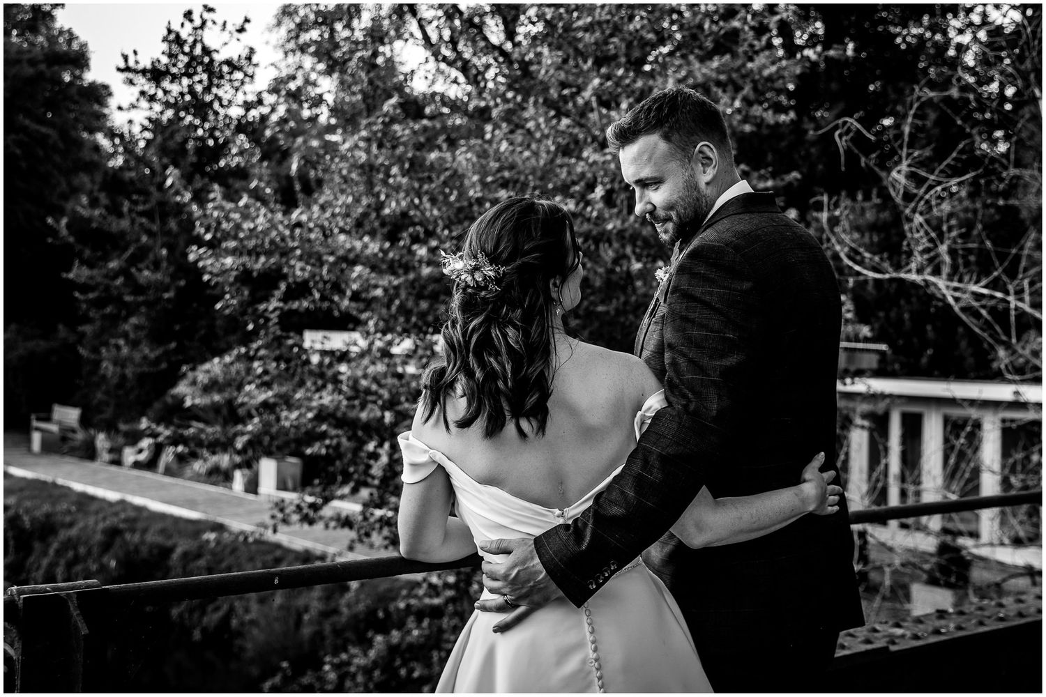 Black and white photo of a bride and groom embracing on a bridge surrounded by lush trees.