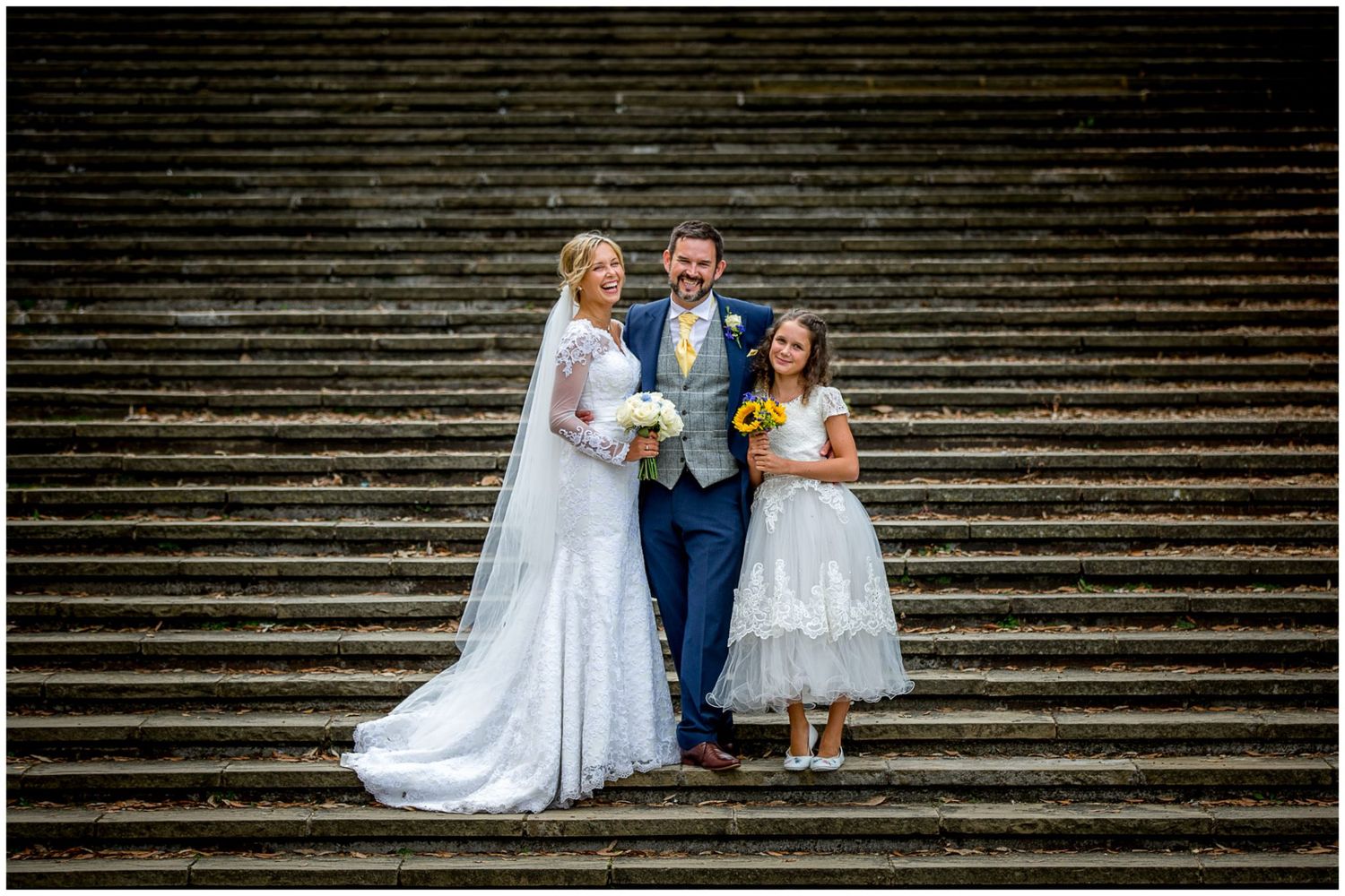 Wedding party members sit together on grand stone steps at a historic venue.