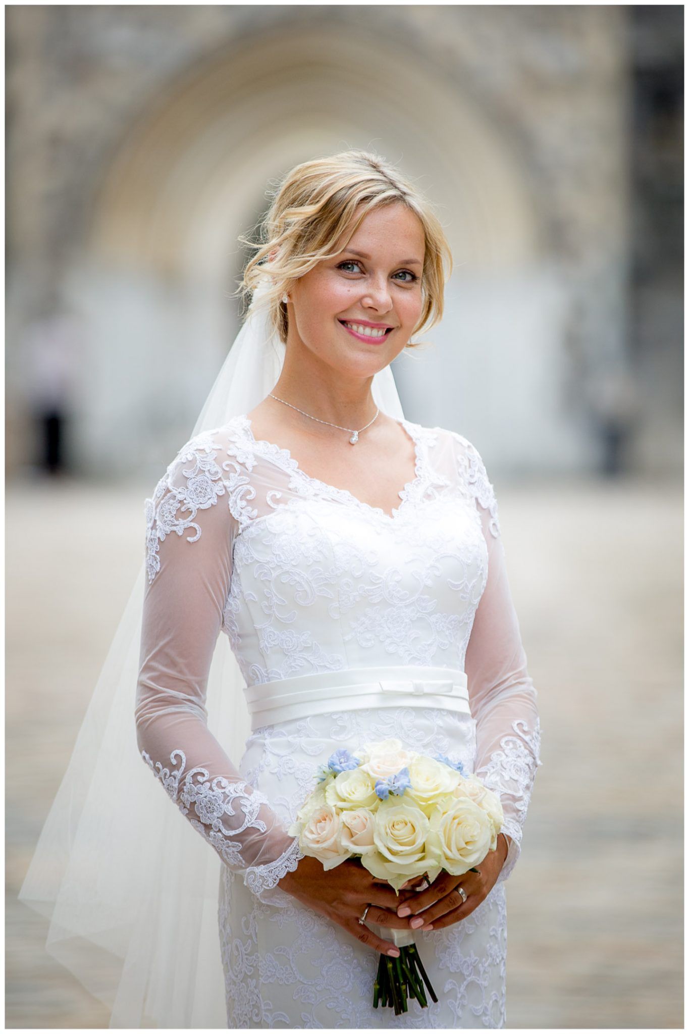 A bride in a white lace long-sleeved wedding gown holds a bouquet of cream roses against a blurred background.