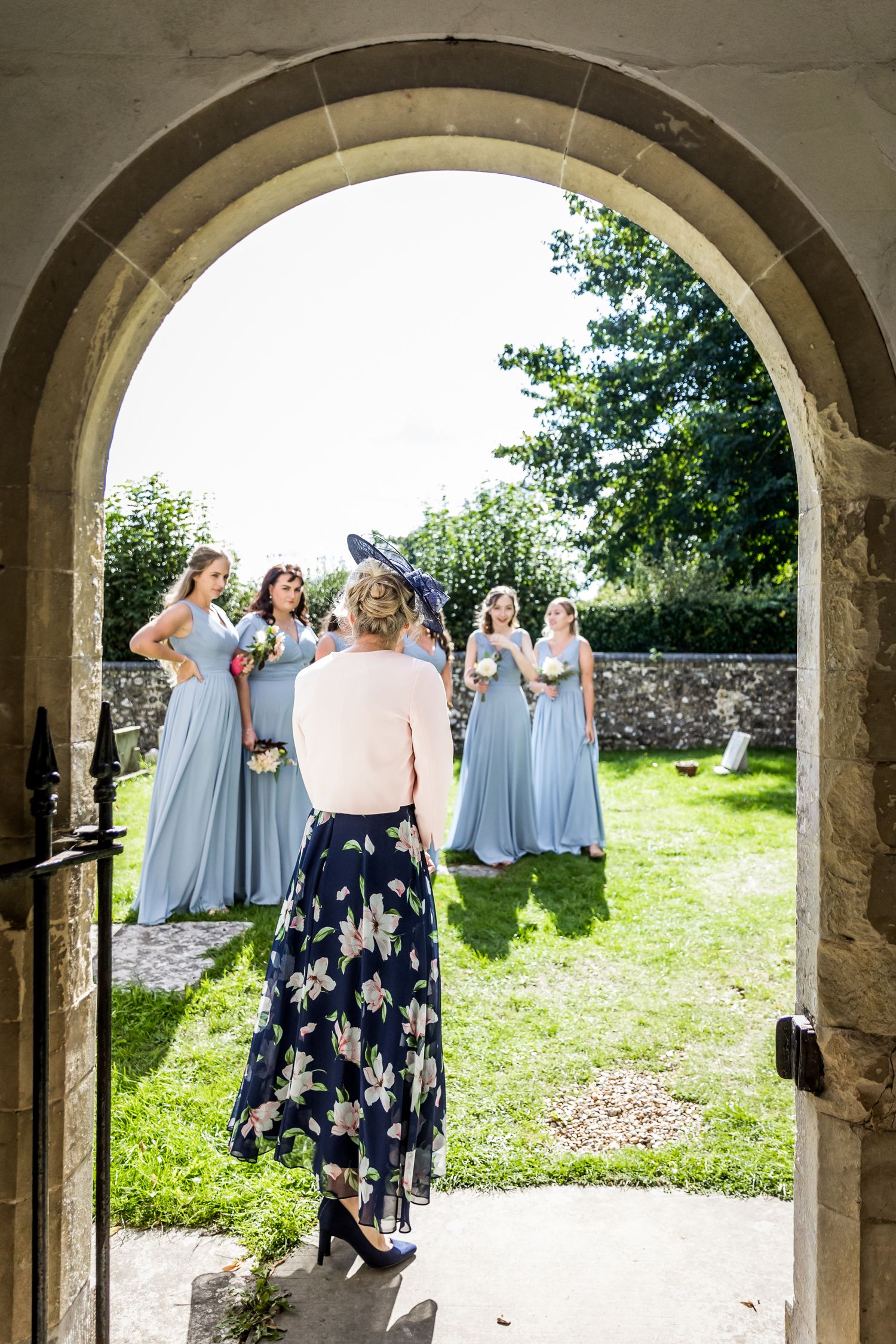 Stone archway frames wedding party gathering on green lawn outside historic church.