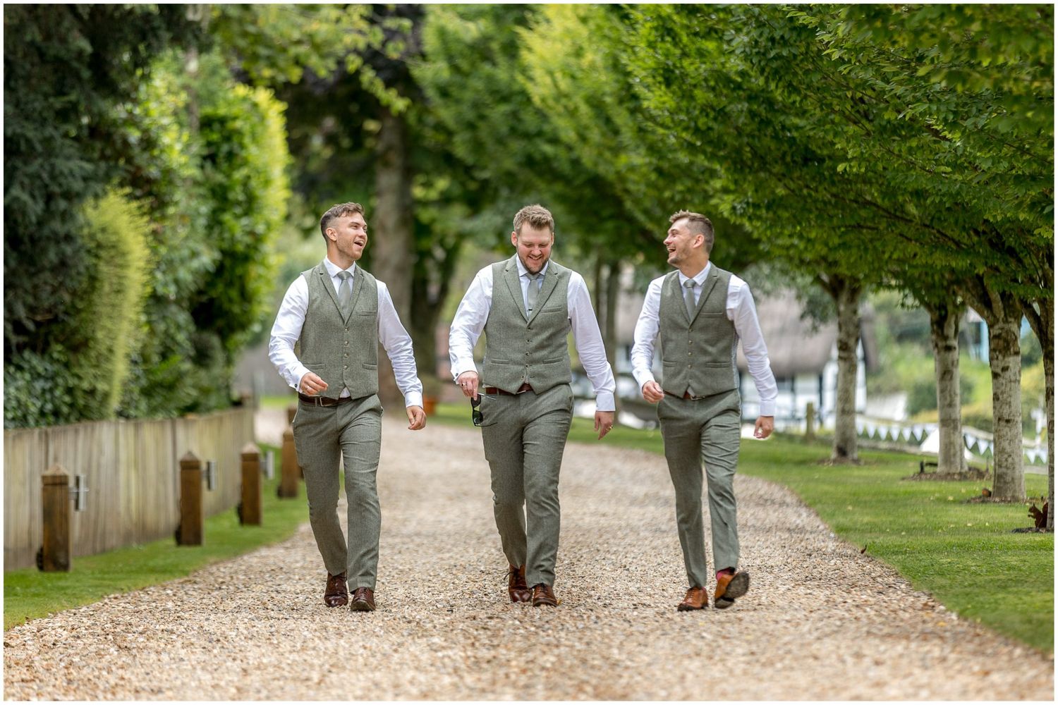 Groomsmen walk together on a tree-lined gravel path wearing matching gray suits.