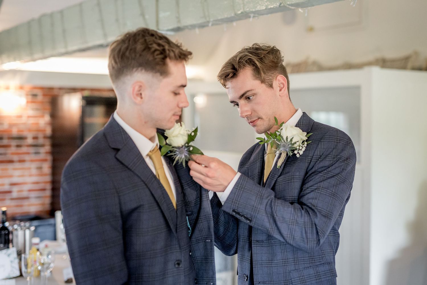Two people in formal wear share a tender moment against white wall and brick backdrop.