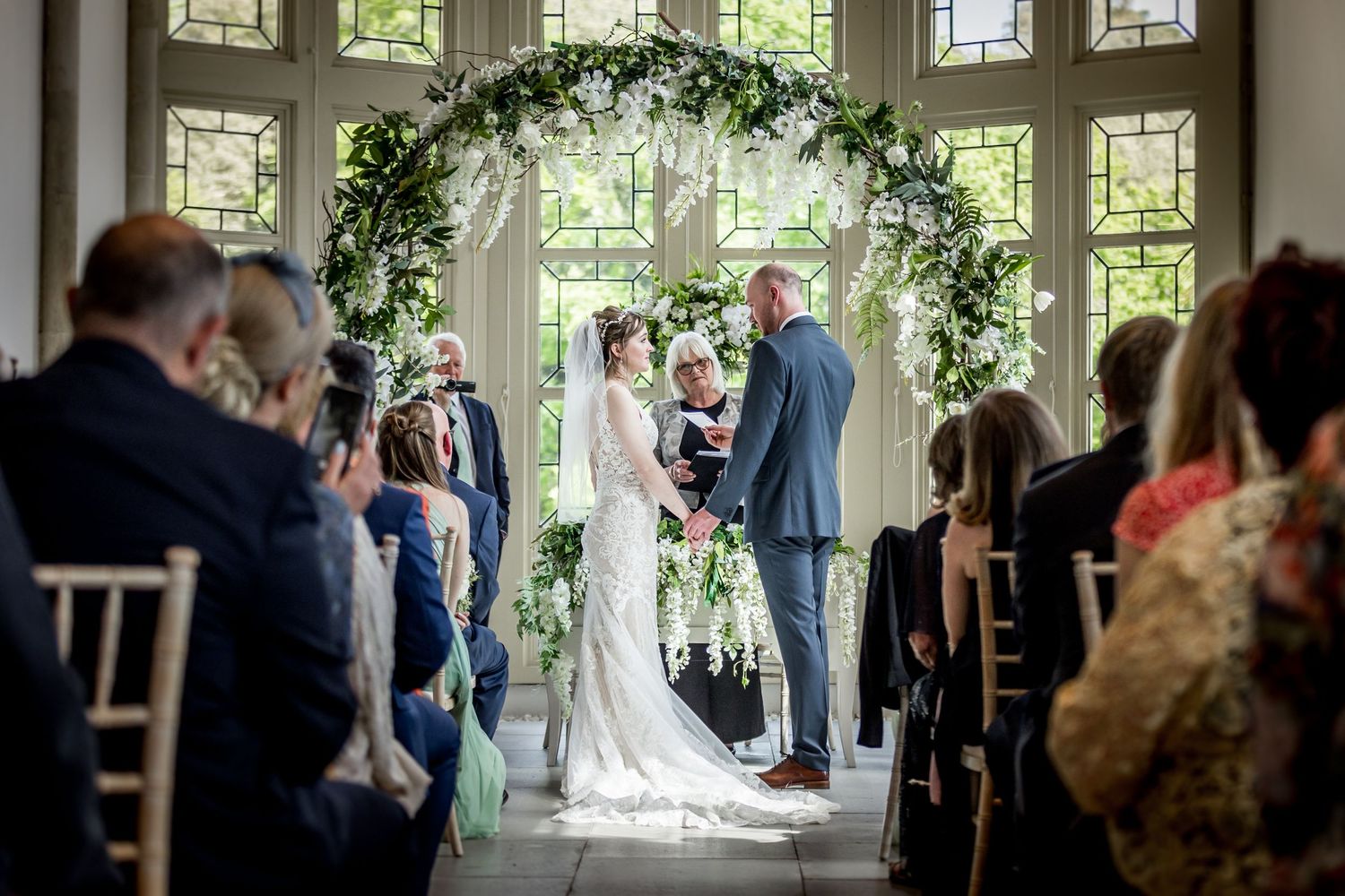 Wedding ceremony takes place under an elegant floral arch with natural light streaming through decorative windows.