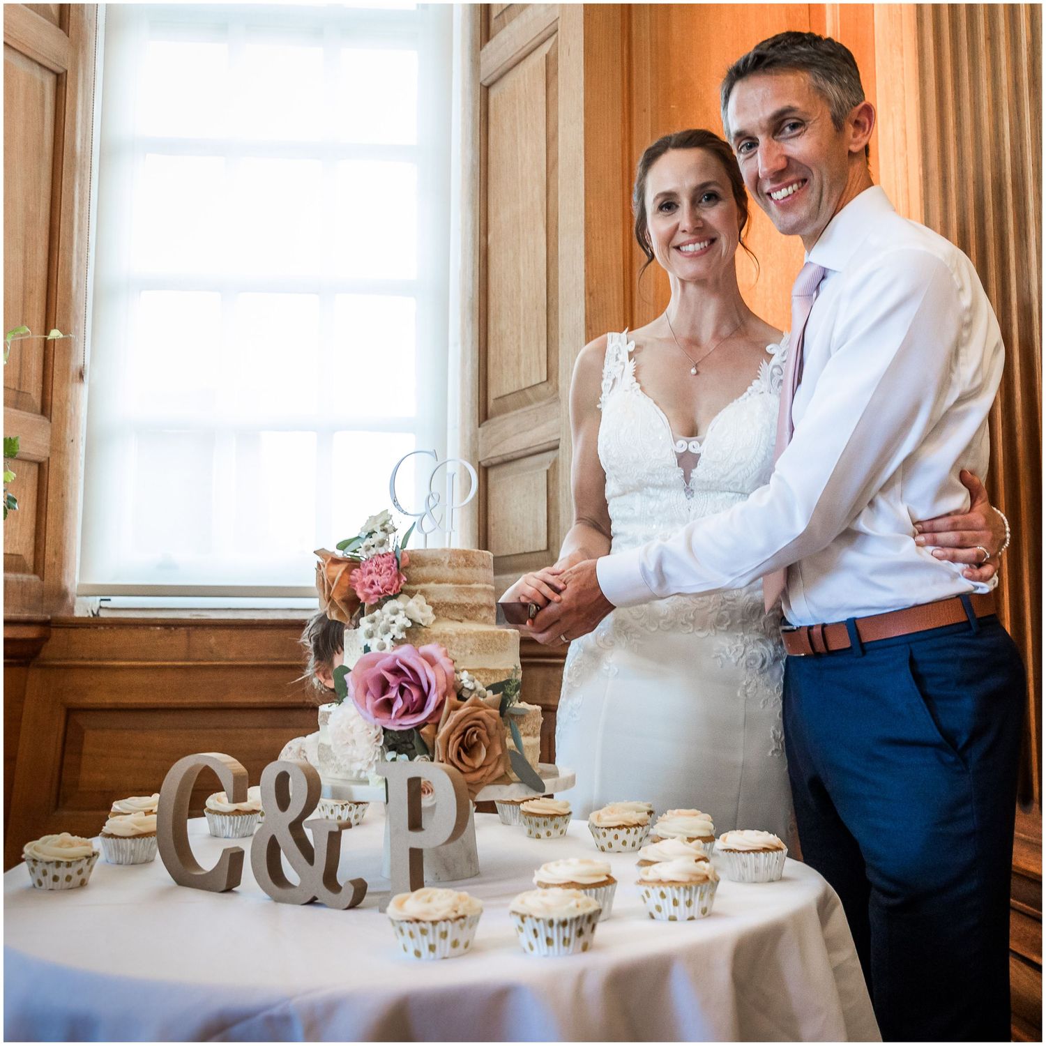 Happy couple cuts their wedding cake decorated with pink flowers and wooden C&P letters with cupcakes.