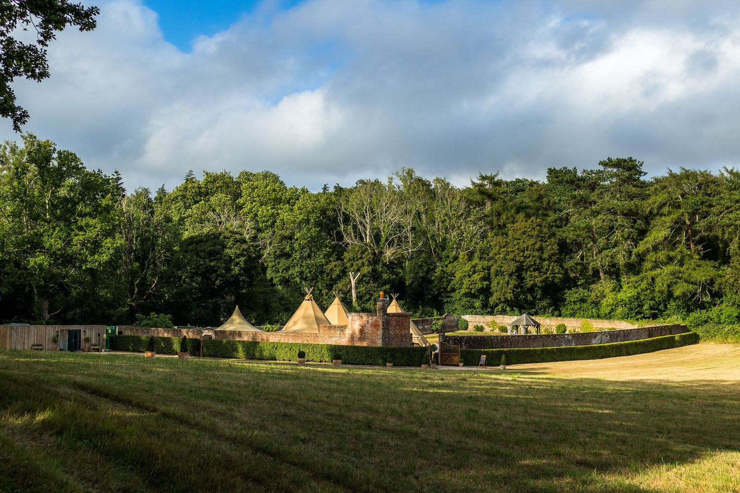 A panoramic view of a grassy meadow with teepee tents and trees in the background under a partly cloudy sky.
