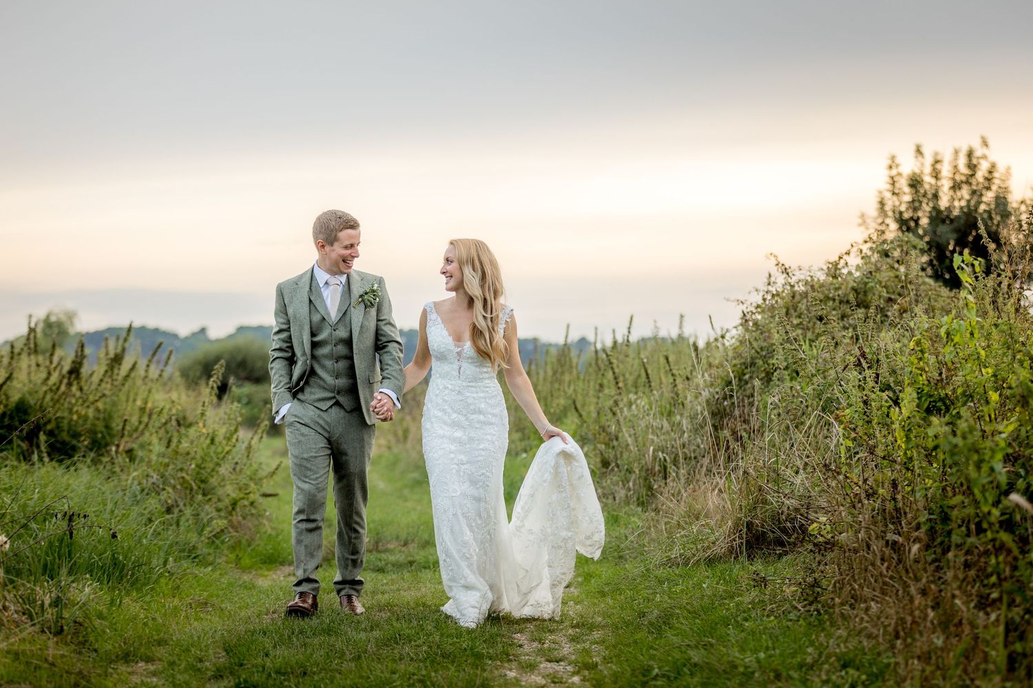 Newlyweds walking hand in hand along a hilltop path at sunset.
