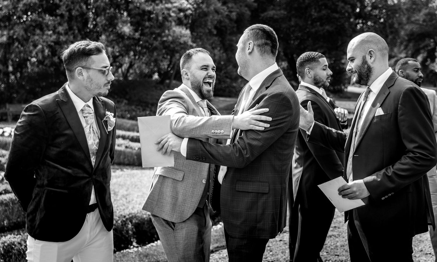 Black and white photo of groomsmen sharing laughs during outdoor wedding celebrations.