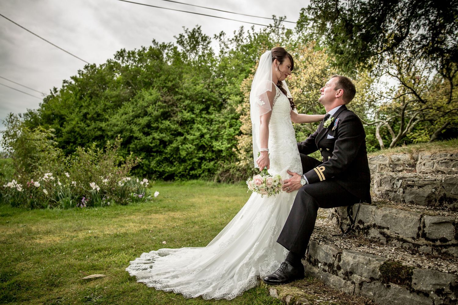 Wedding couple poses on rustic stone wall surrounded by lush greenery.