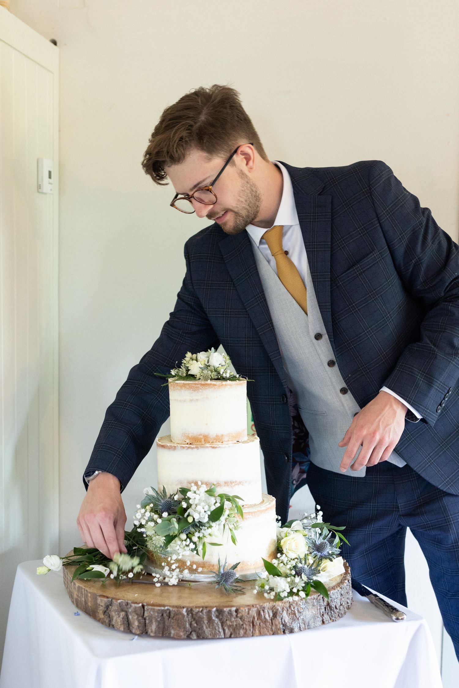 Person in navy suit arranges a rustic wedding cake on wooden stand.