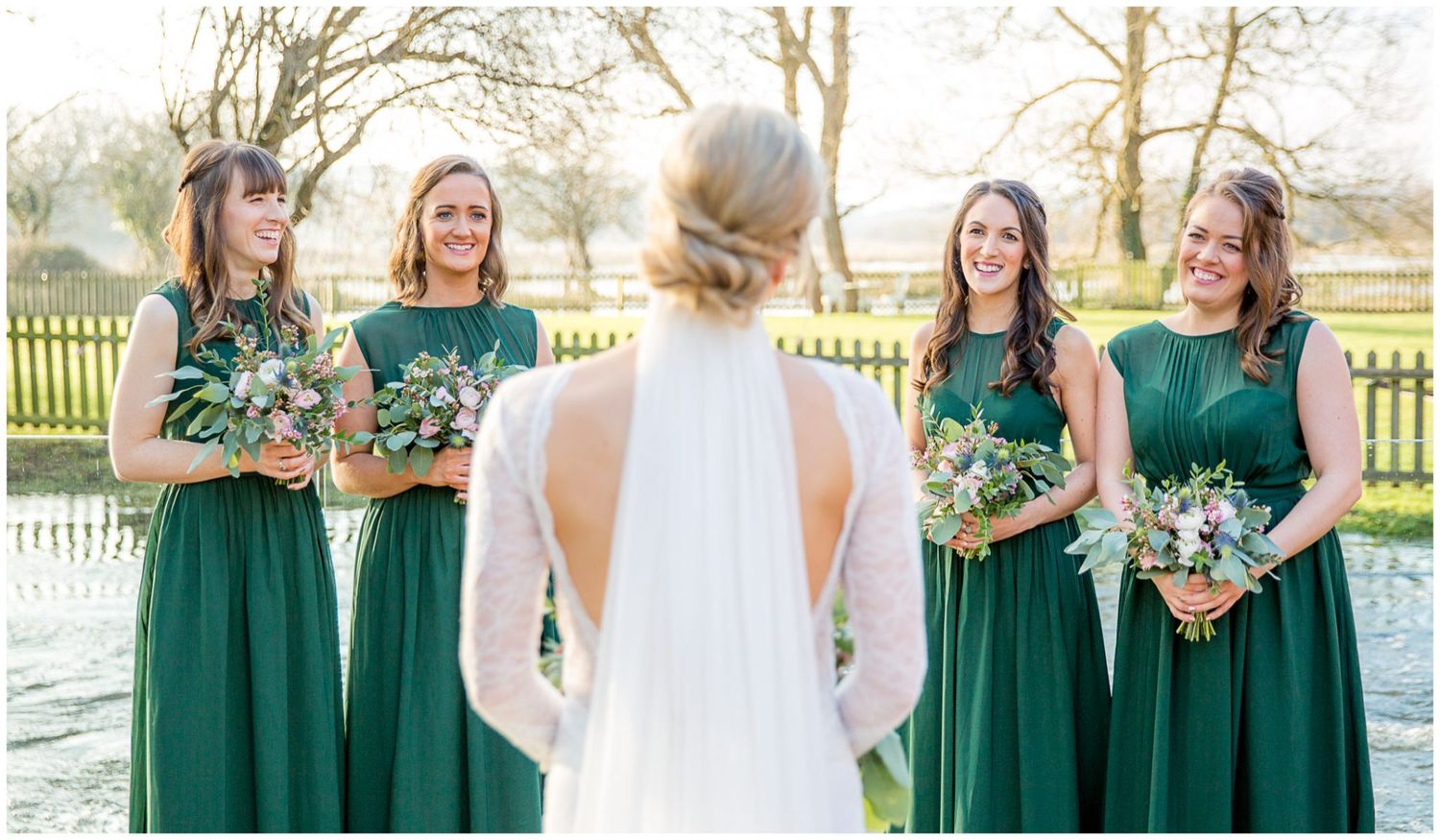Bridesmaids in emerald green dresses stand together holding bouquets outdoors.