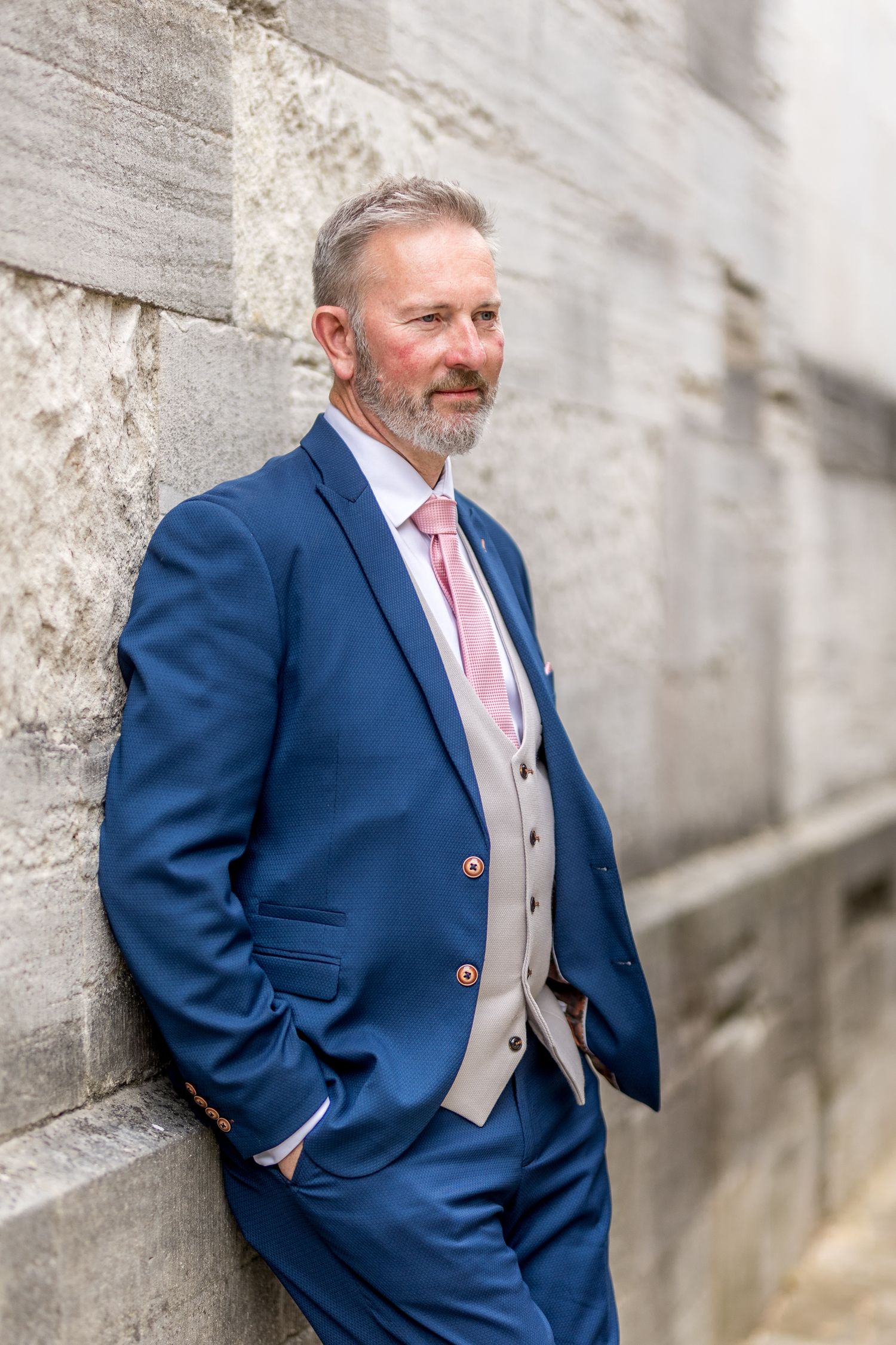A groom in a navy suit and pink tie poses against a light stone wall.