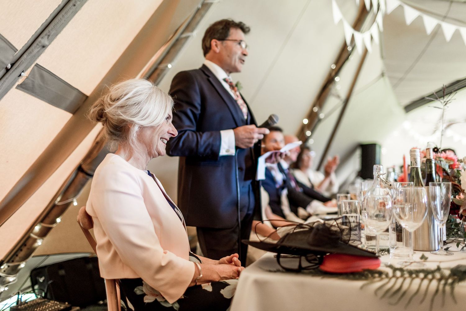 Wedding guests laughing and enjoying speeches during a reception dinner under white bunting decorations.