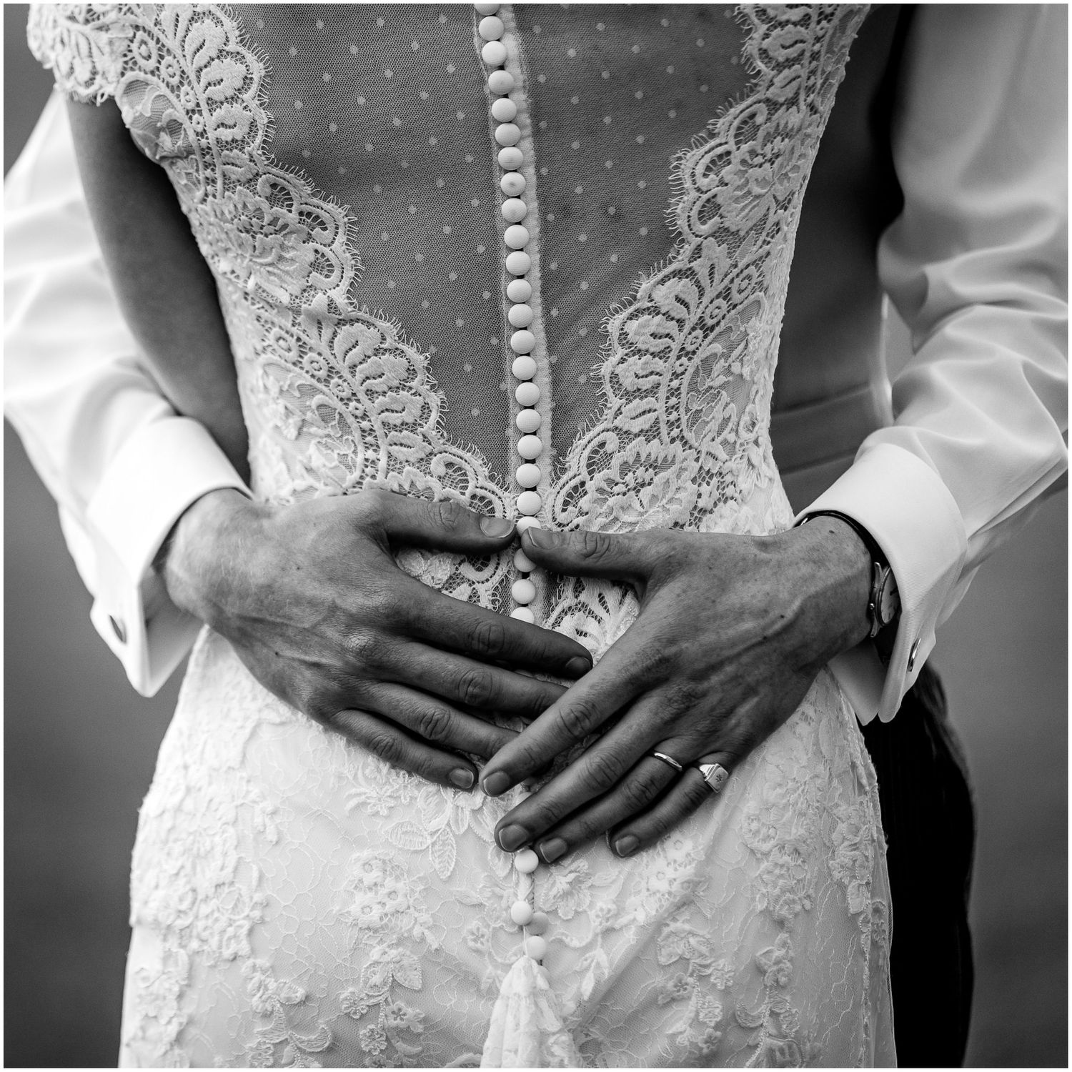 Close-up black and white detail of lace wedding dress with hands showing wedding ring.