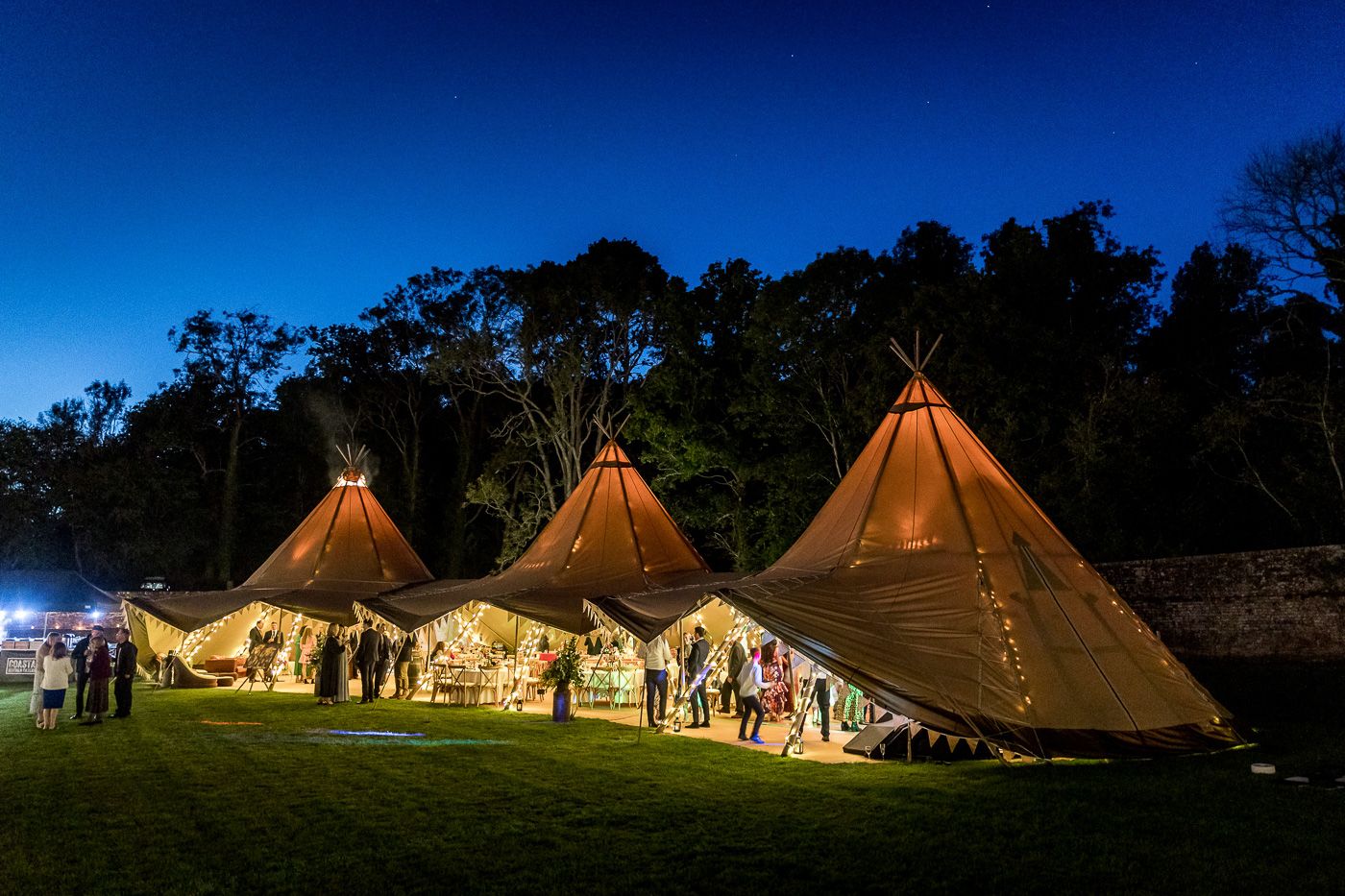 Three illuminated tipi tents glowing at night during an outdoor wedding reception.