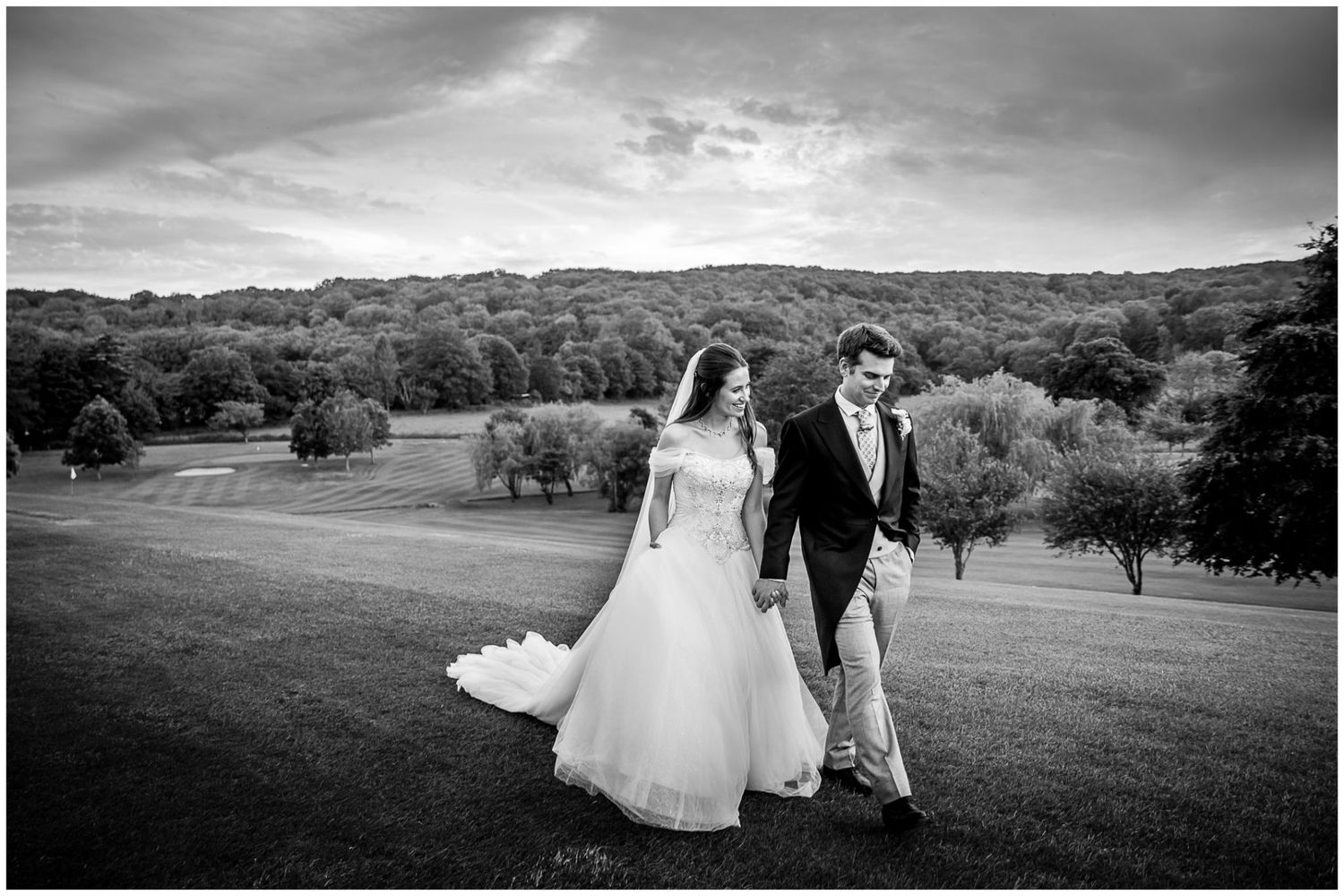 A black and white wedding photo showing a couple walking hand in hand across a scenic hillside landscape.