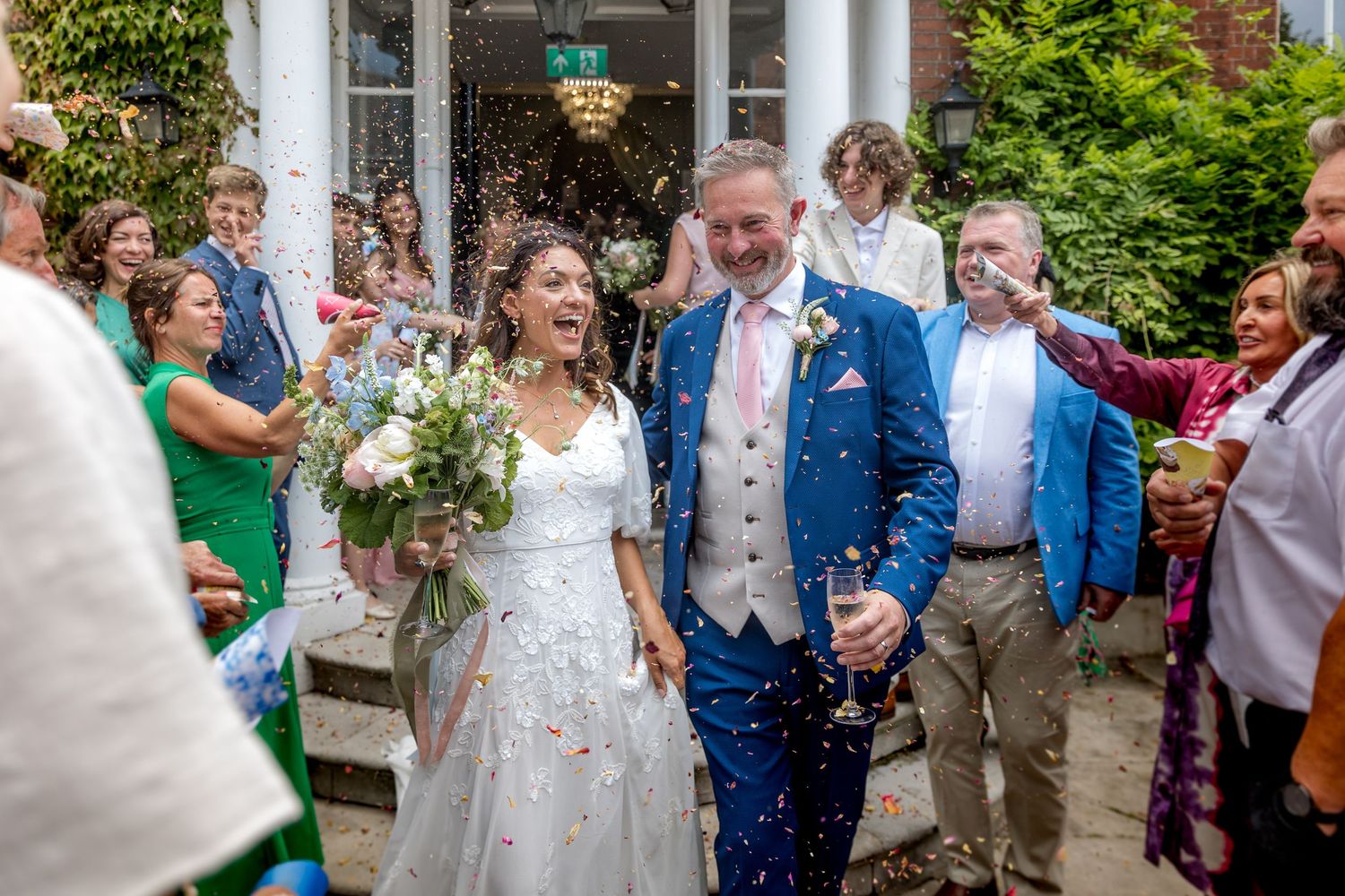 Newlyweds celebrating with guests throwing confetti outside venue entrance.