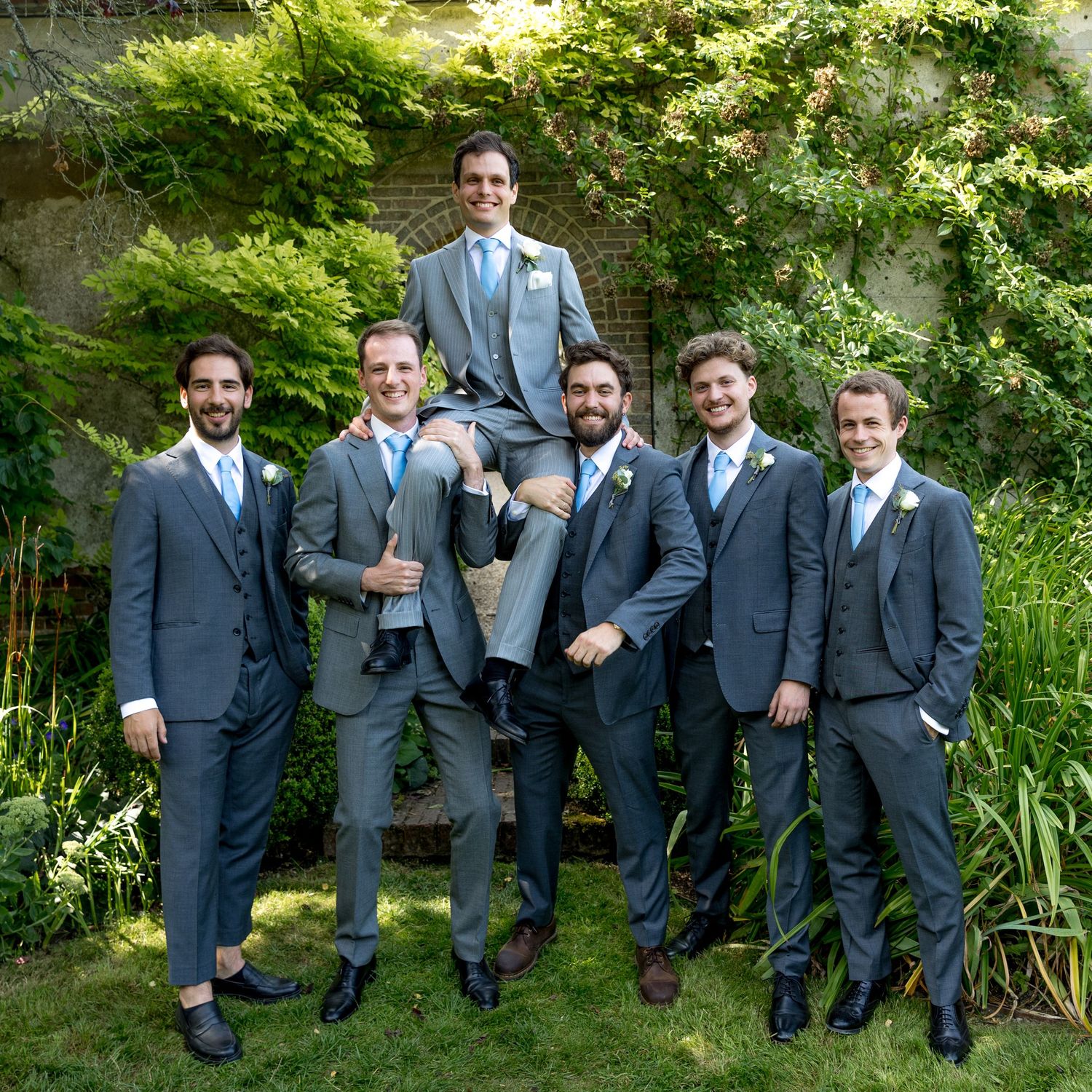 Groomsmen in matching grey suits pose together in the walled gardens of Houghton Lodge during the group photograph session