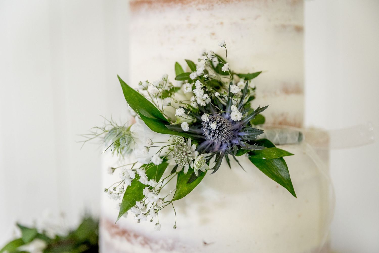 Elegant white wedding cake decorated with delicate blue thistle and fresh greenery against light background.