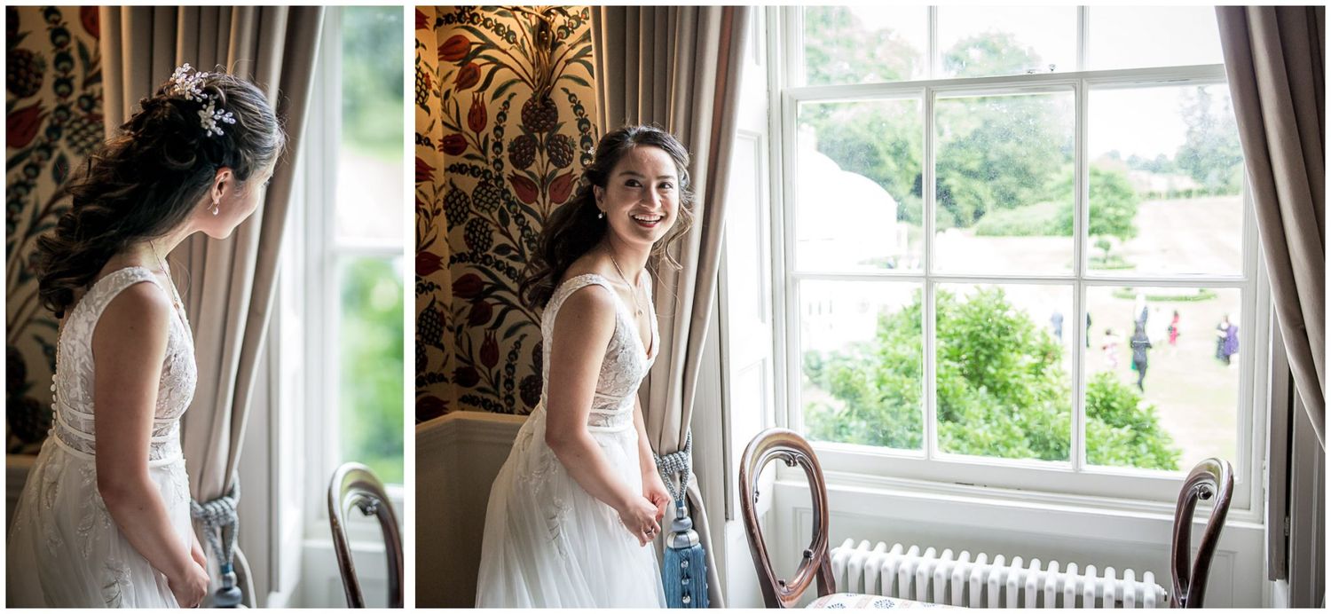 Bride in white dress poses elegantly by window with natural light streaming in during wedding preparations.
