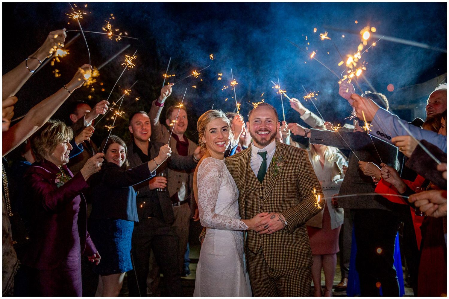 Wedding guests hold sparklers creating magical nighttime celebration under dark blue sky.
