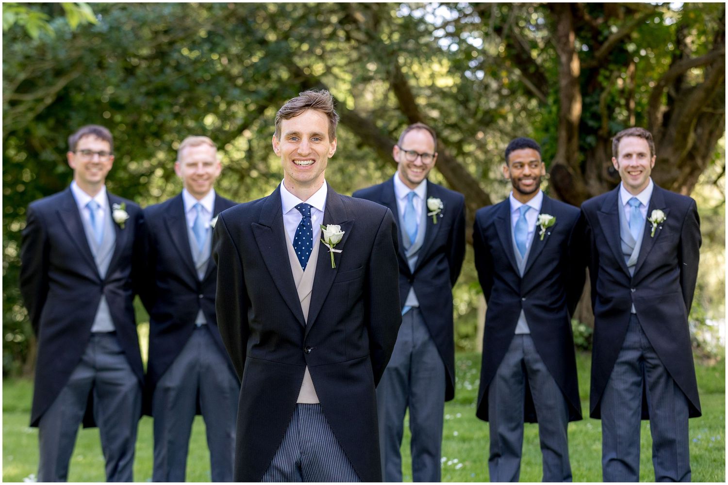 Groomsmen in dark suits pose together on green lawn.