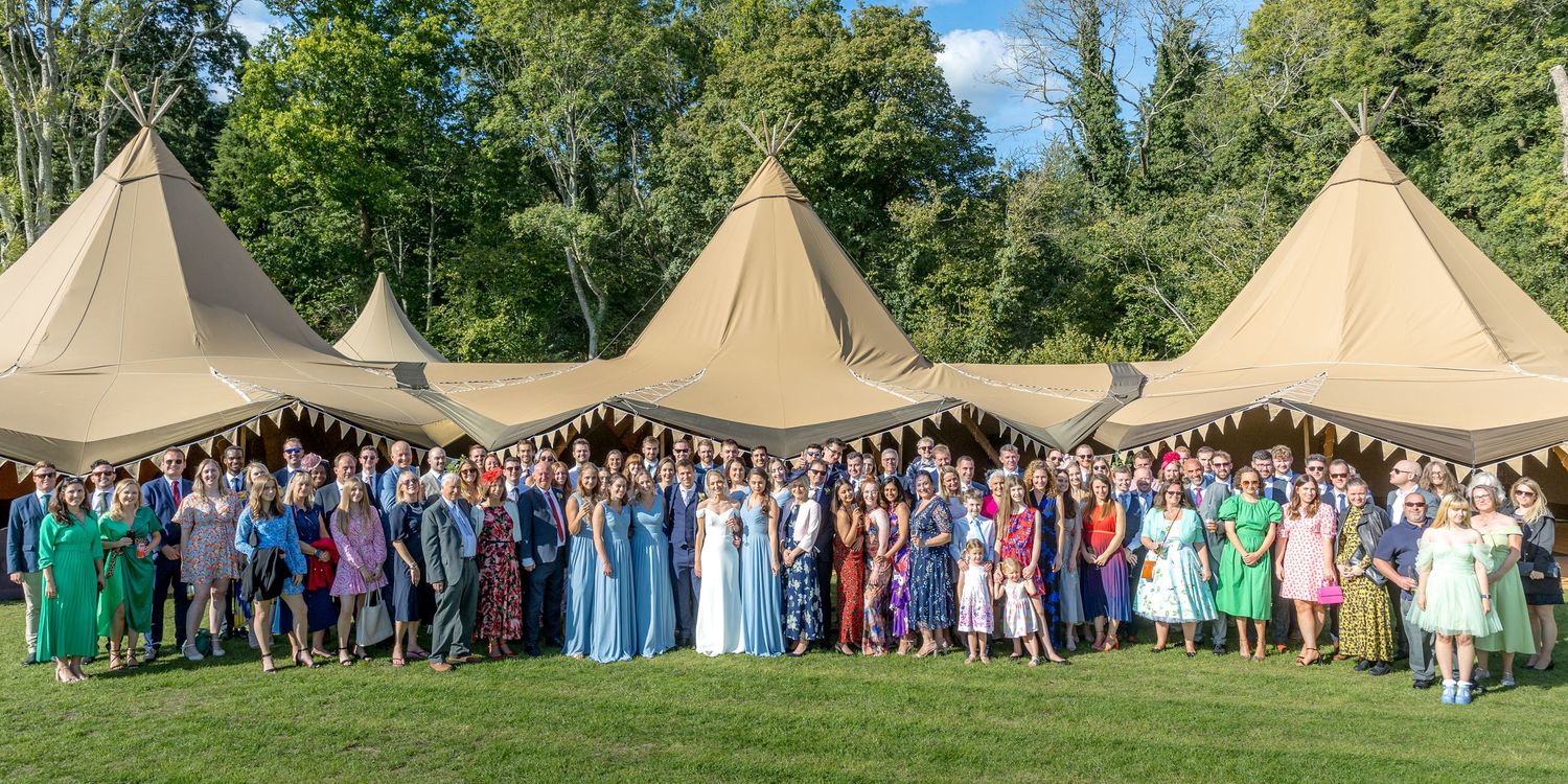 Large group photo of wedding guests posing in front of tipi tents on a sunny day.