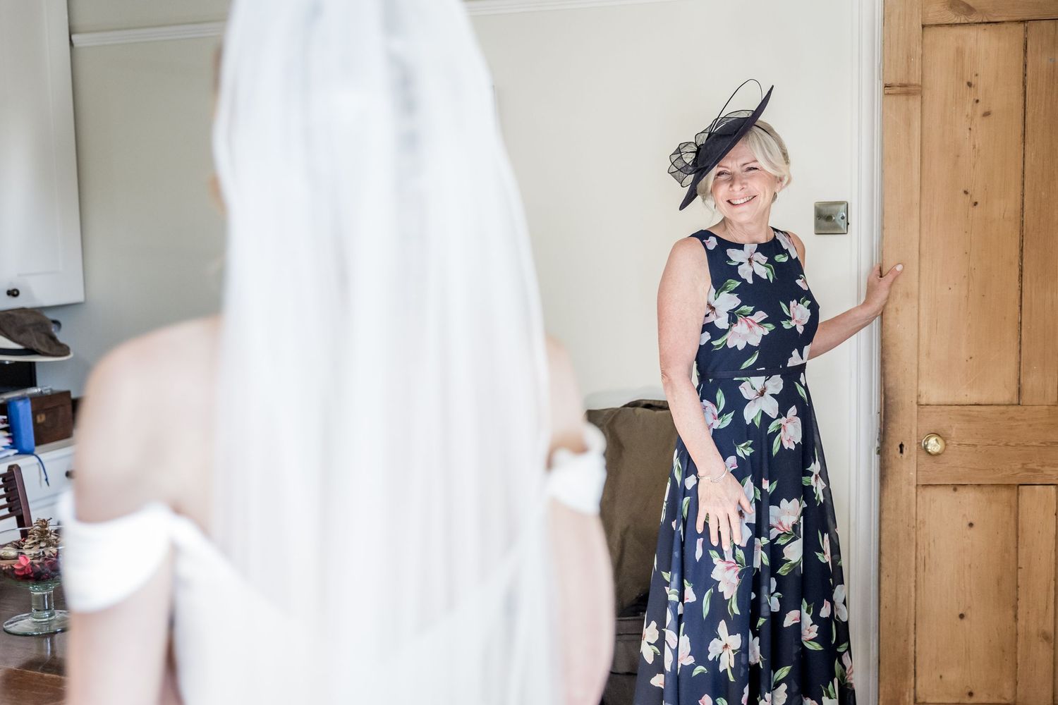 A woman in a navy floral dress and fascinator reacts emotionally while looking at a bride in a white gown.