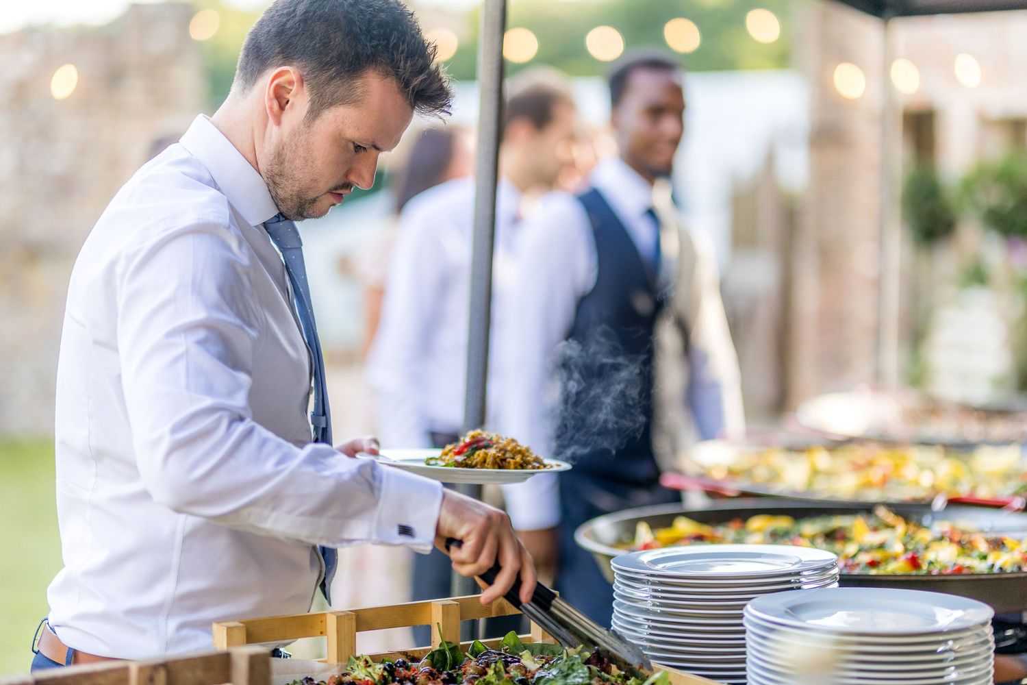 Catering staff member in white shirt serving food at an outdoor buffet station.