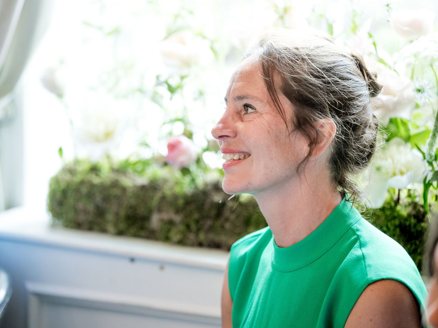 A person in a bright green shirt smiles while looking to the side in a sunlit room with plants.