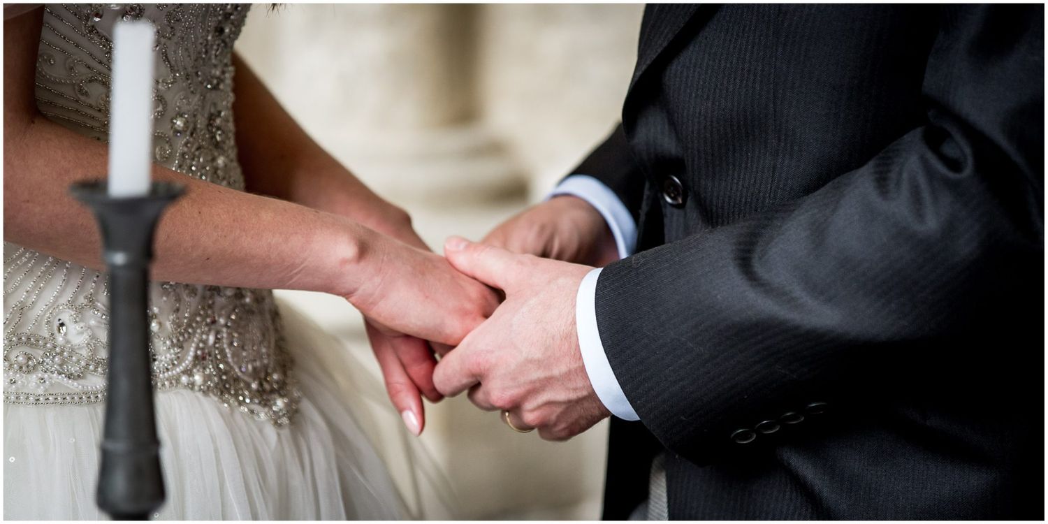 Close-up of bride and groom holding hands during wedding ceremony with white dress and black suit visible.