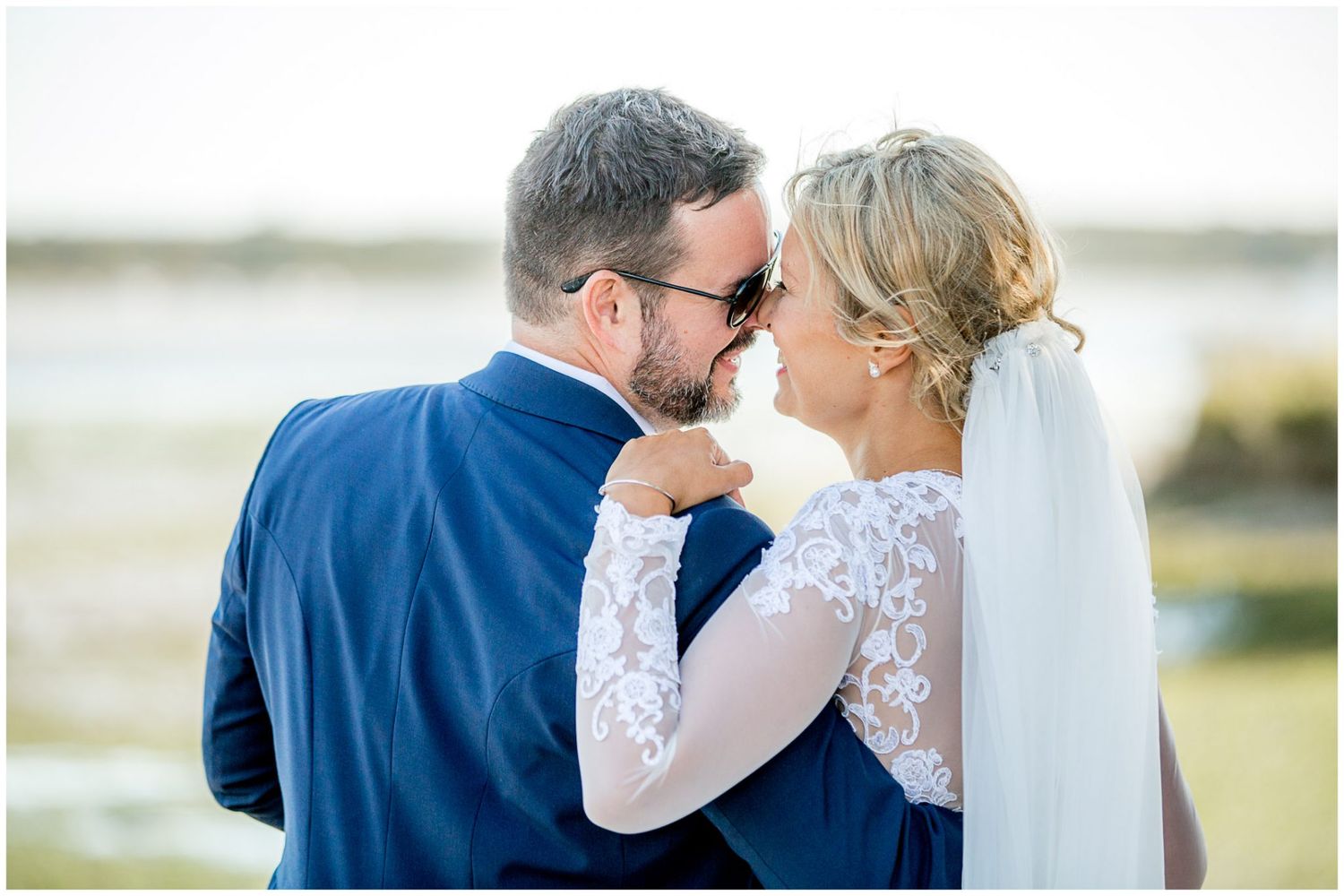 A bride in a long sleeve lace dress embraces her groom in a navy suit during an outdoor coastal wedding.