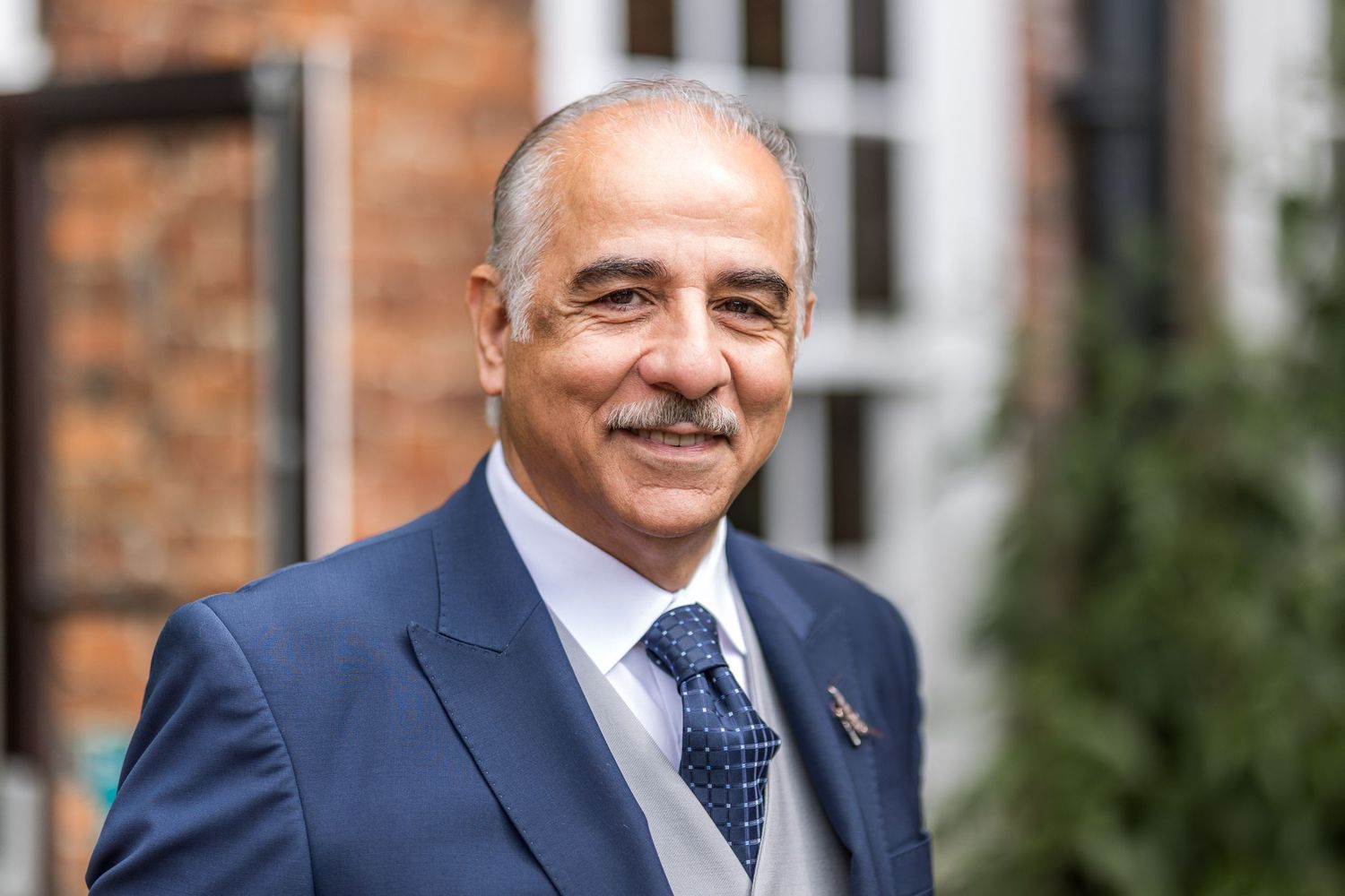 Gentleman in navy suit smiling outdoors against brick building.